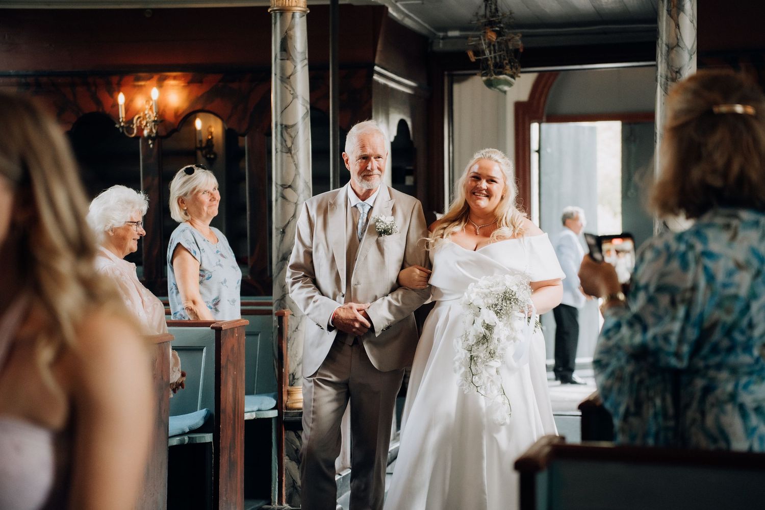 Smiling bride in white gown walks down church aisle arm-in-arm with escort while guests applaud and photograph.