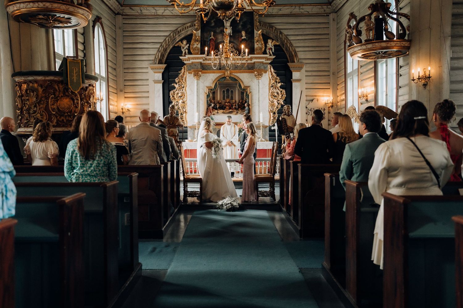 Wide shot of wedding ceremony in elegant historic church with guests seated in wooden pews and ornate altar.