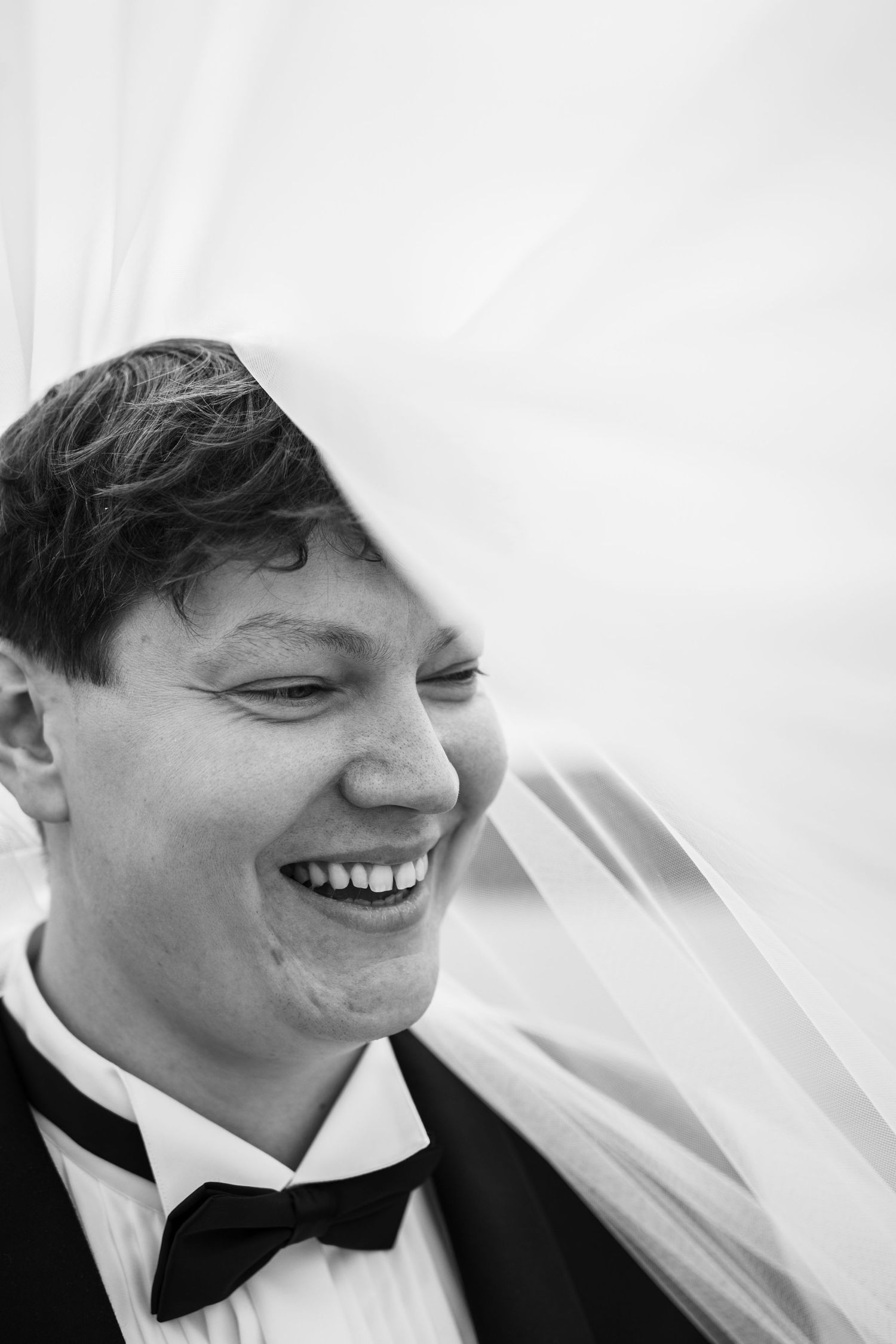 Groom laughing joyfully under bride's veil in black and white wedding portrait.