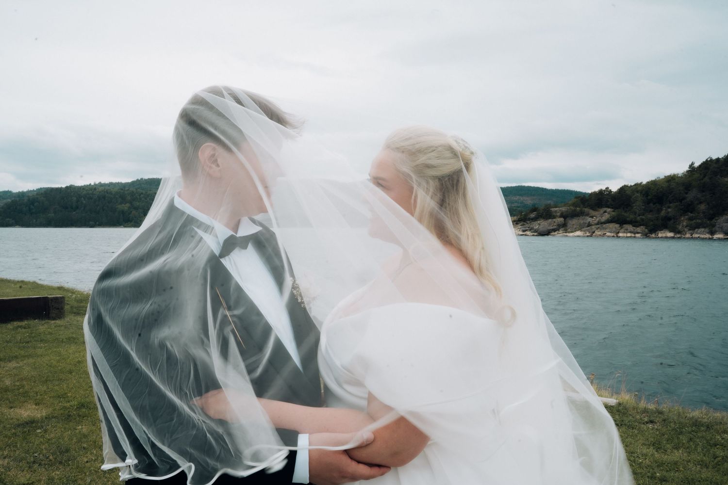 Bride and groom embracing under a flowing veil with a scenic lake and green hills in background.