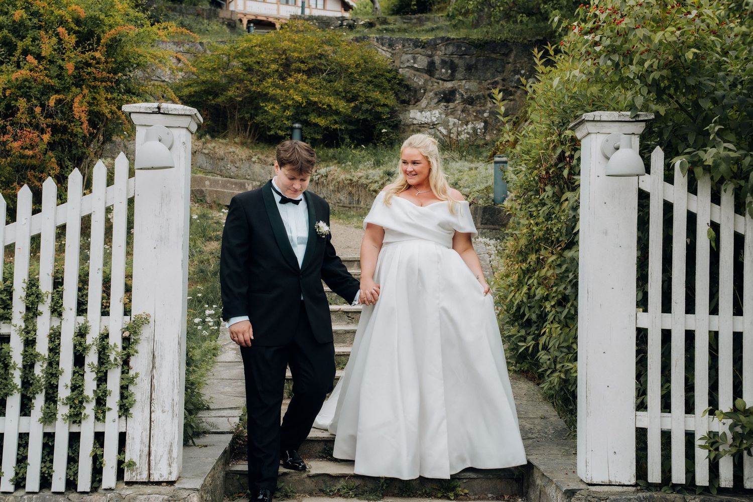 Bride in white ball gown and groom in black tuxedo walking through a white garden gate on steps.