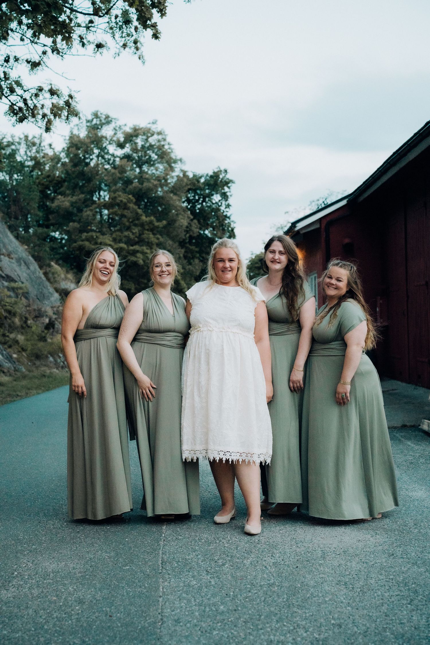 Bride in short white lace dress posing with four bridesmaids in sage green multiway gowns outdoors.