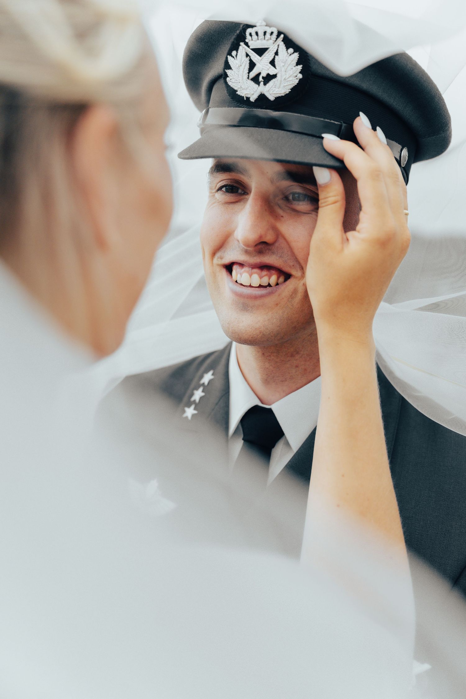 Smilende brudgom i militær paradeuniform og uniformslue mens bruden justerer luen hans under sitt flagrende hvite slør.