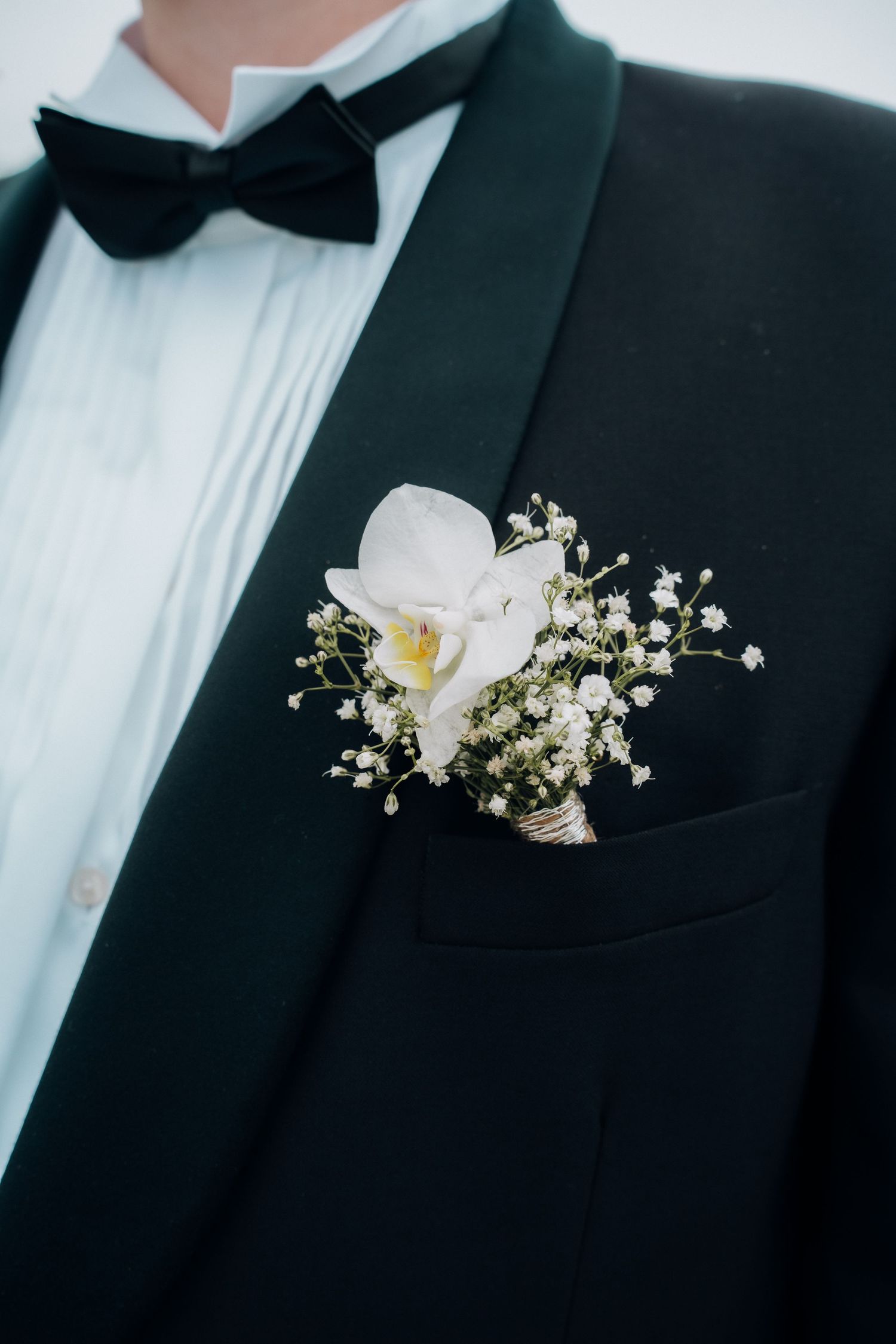 Close-up of groom's black tuxedo lapel with white orchid and baby's breath boutonniere and bow tie.