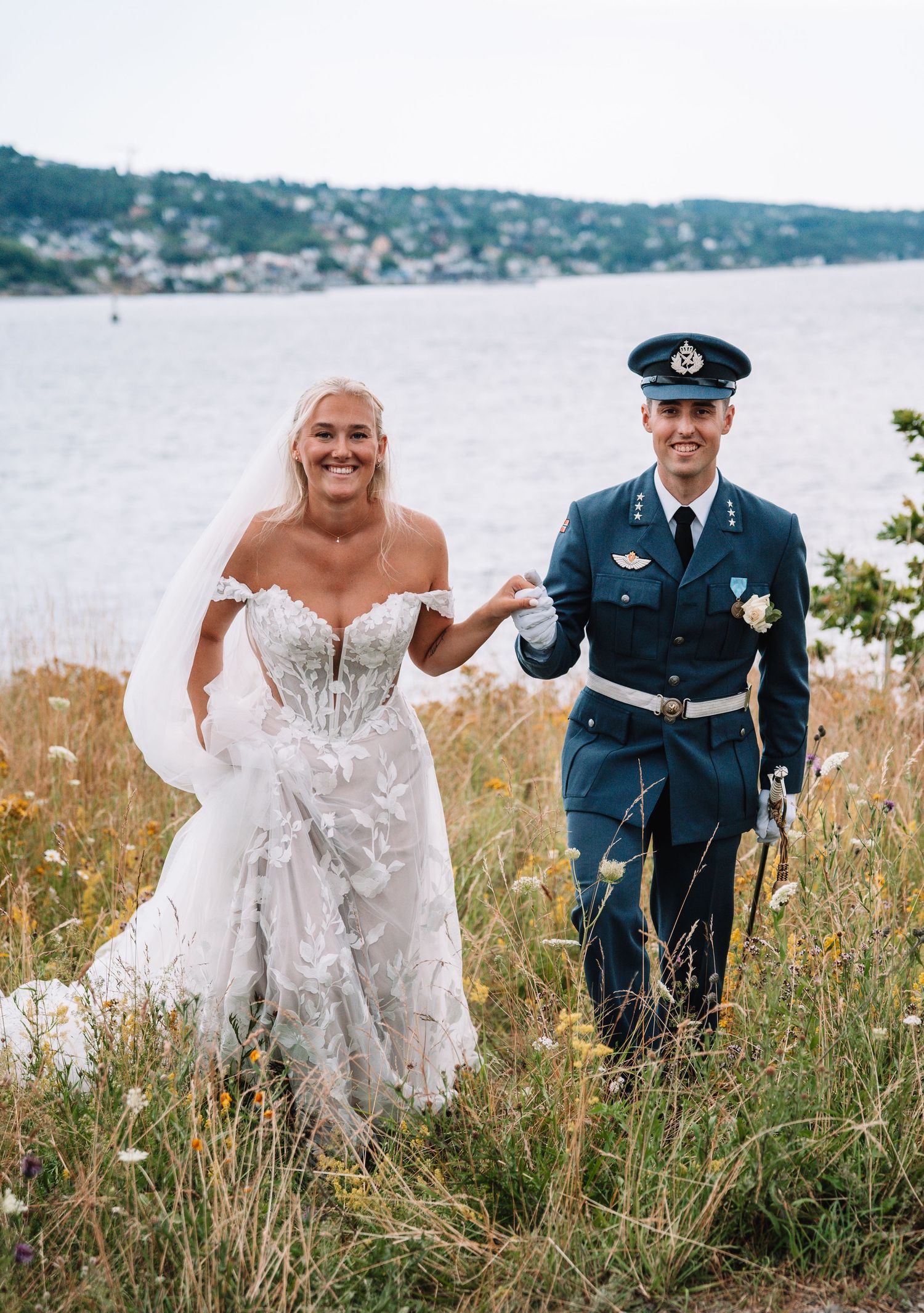 Bride and groom in military uniform walk hand in hand through a wildflower field overlooking a fjord.