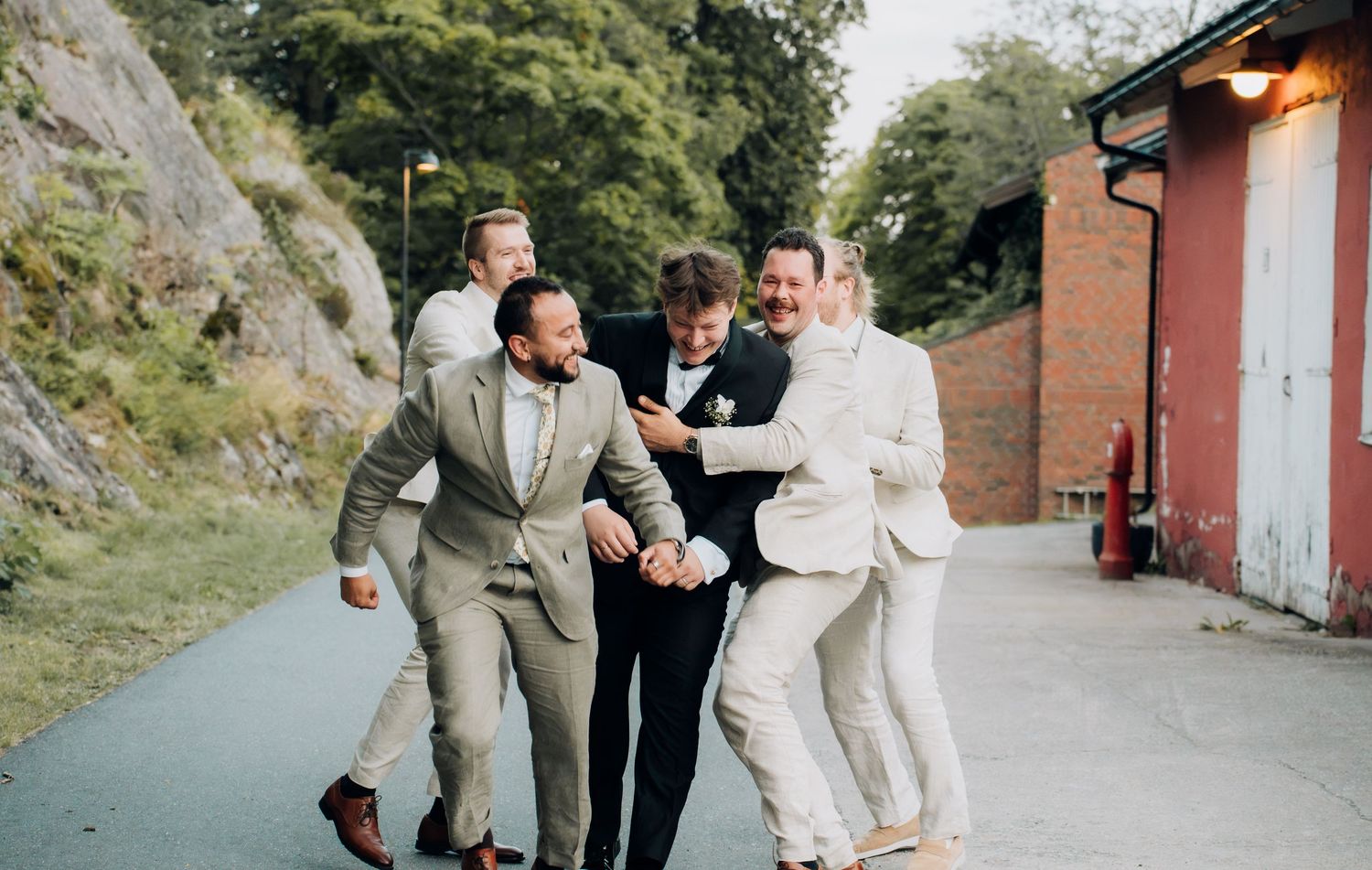 Four groomsmen in linen suits playfully celebrate together along an outdoor wedding venue path.