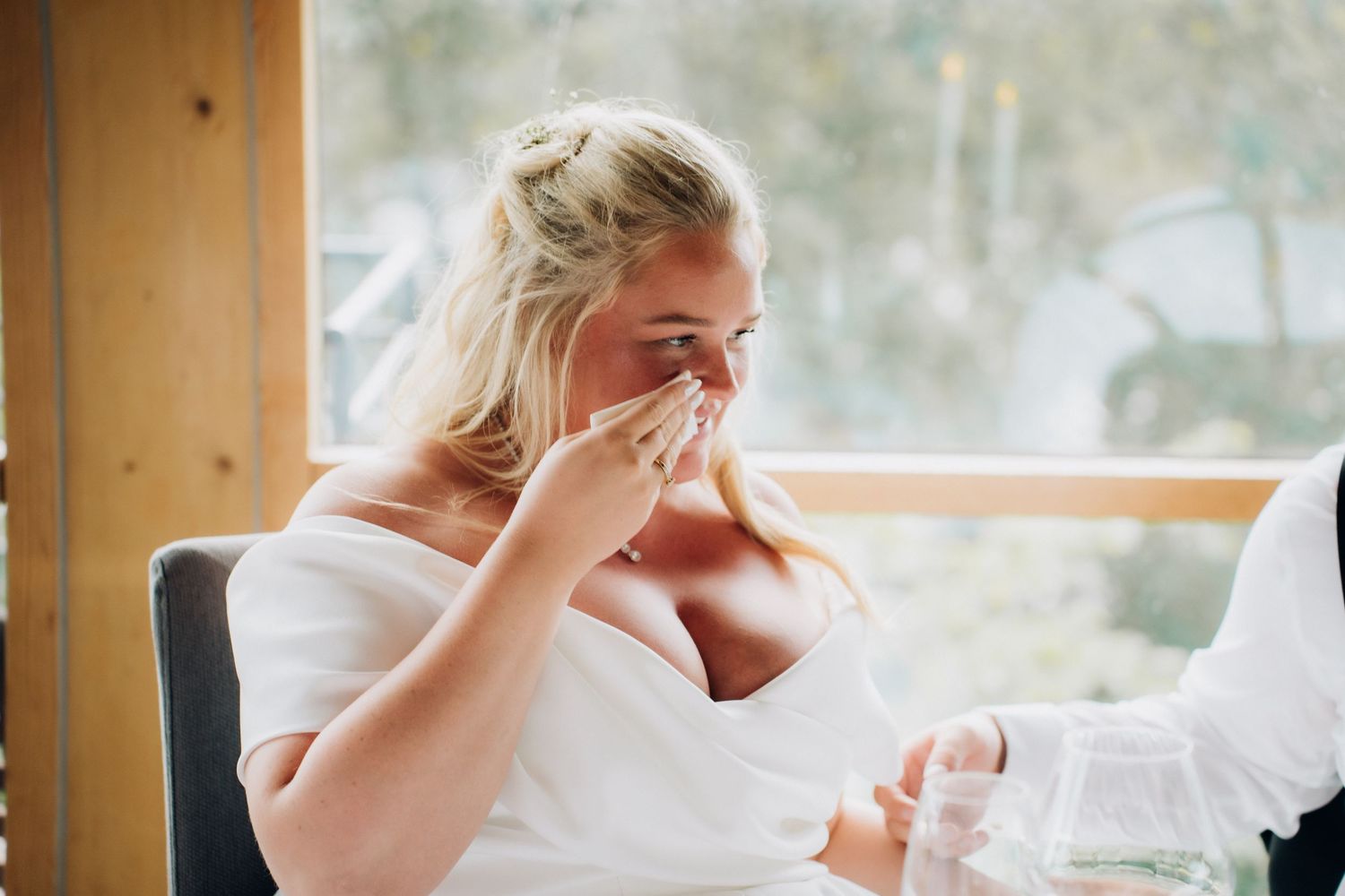 Emotional bride in white off-shoulder dress dabbing tears with tissue during wedding ceremony.