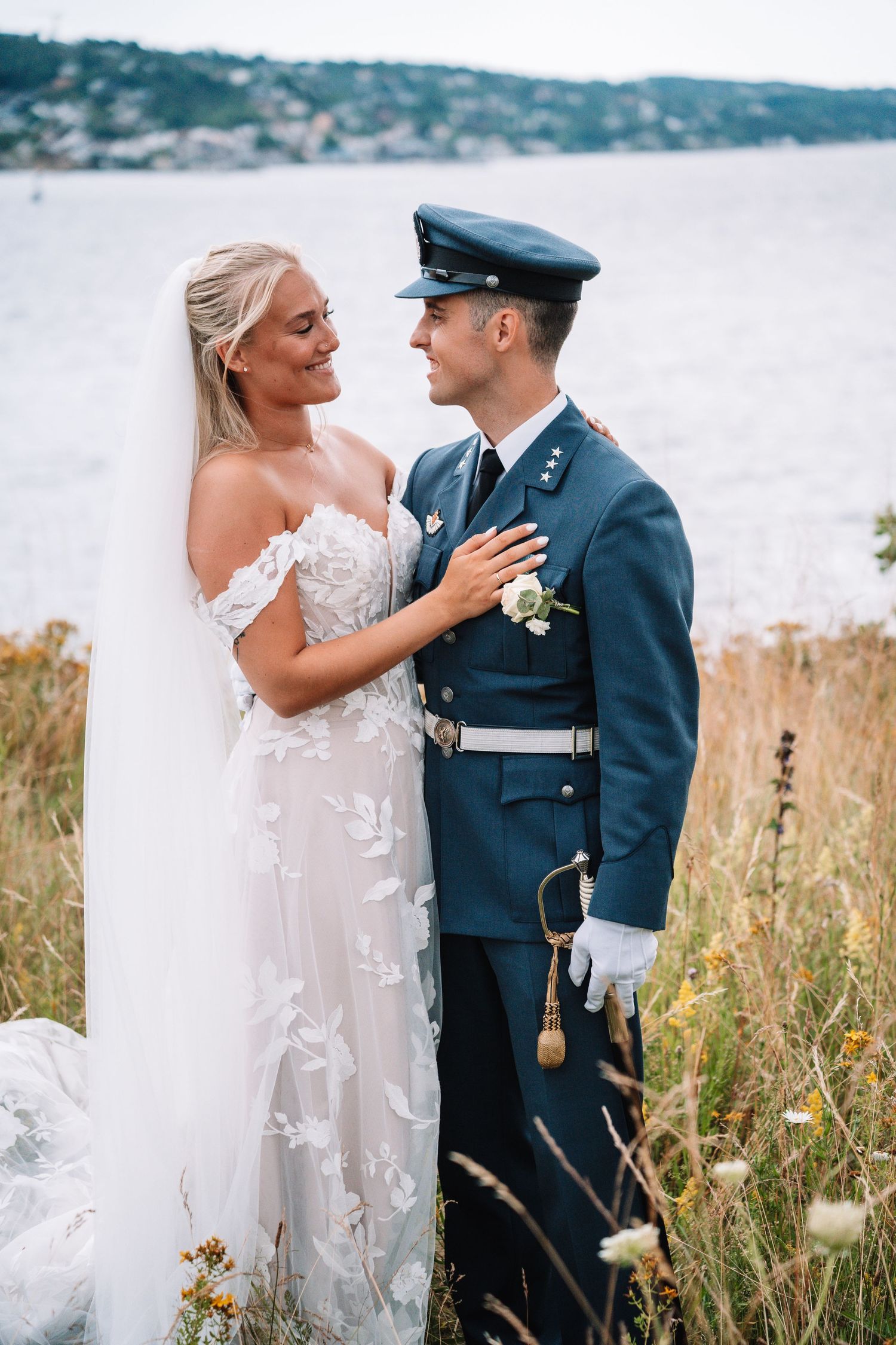 Brud i blomstrete blondekjole og brudgom i blå militæruniform omfavner hverandre i en gyllen kysteng.