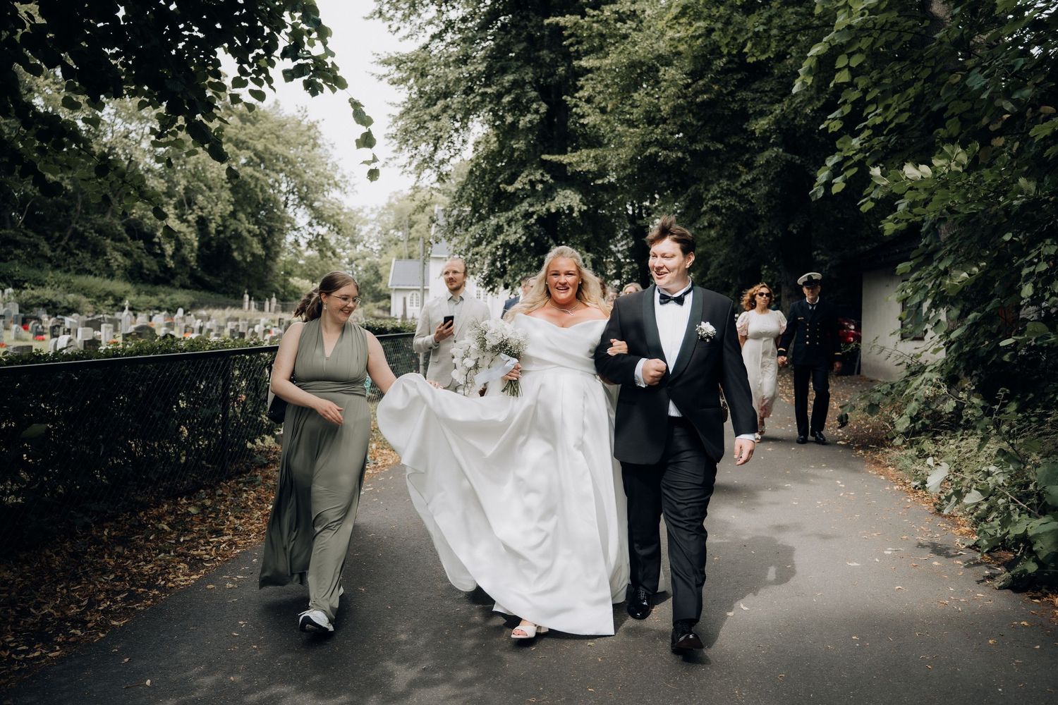 Bride and groom walking joyfully with wedding party through a tree-lined pathway.