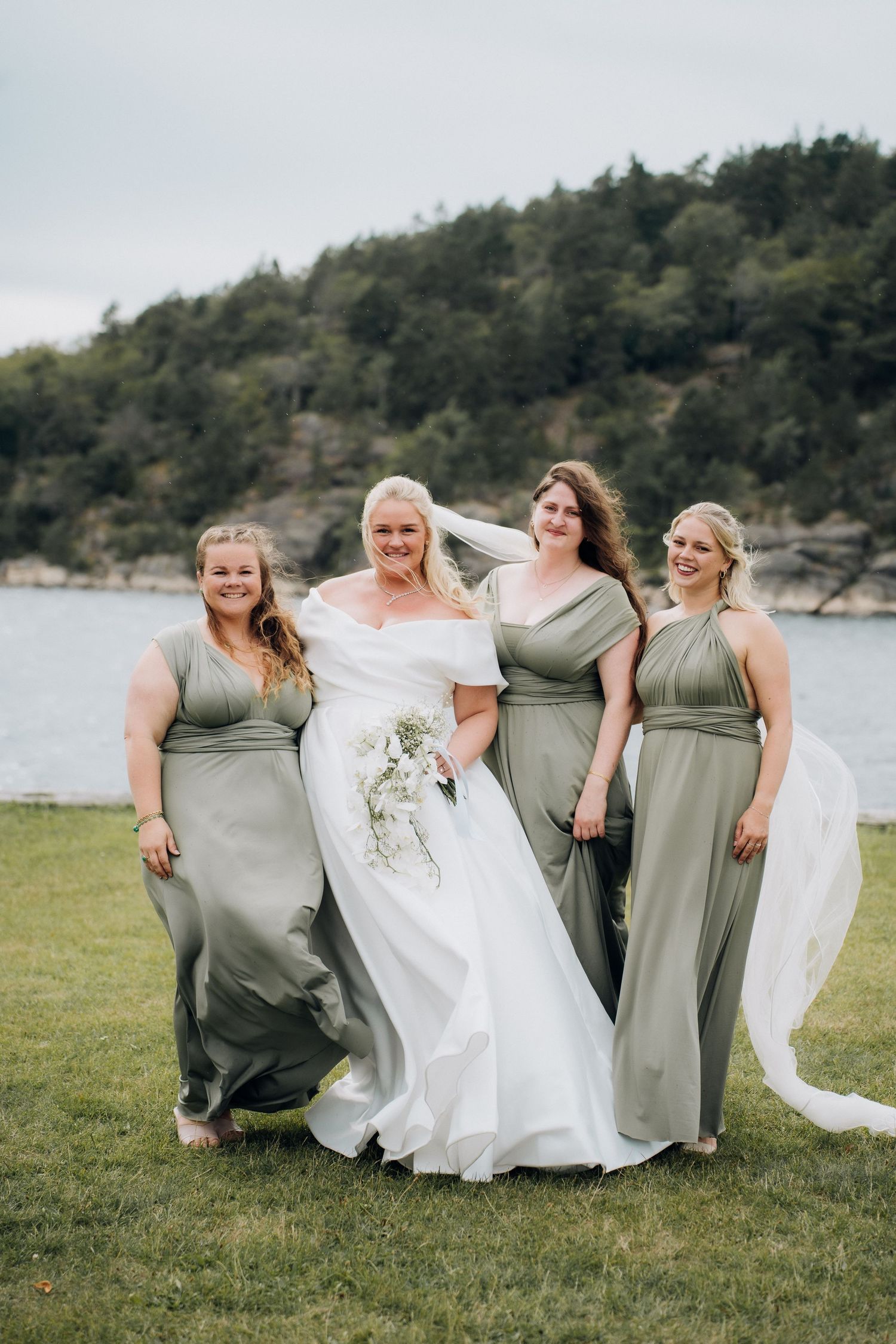 Bride in white gown poses with three bridesmaids in sage green dresses by waterfront.