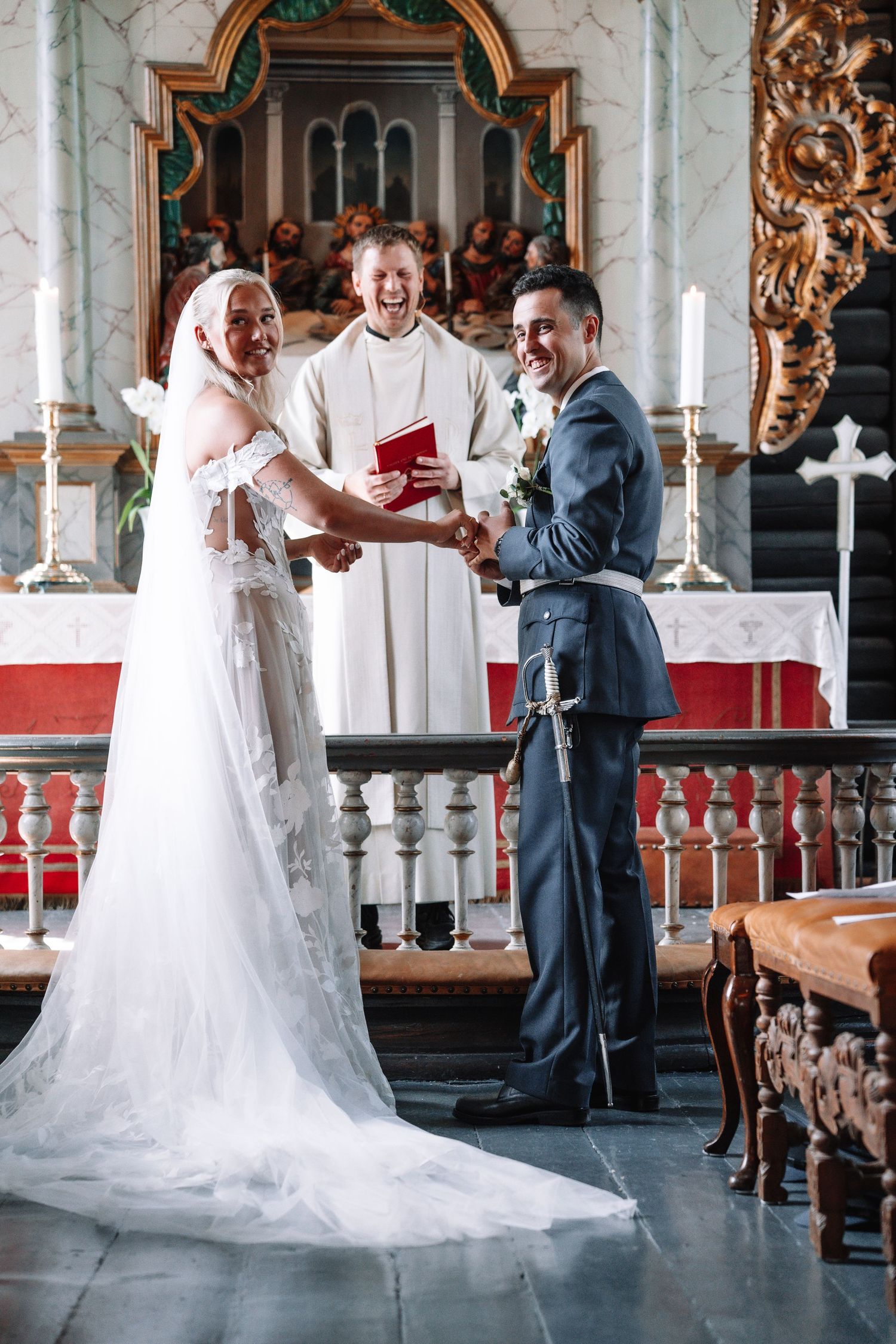 Brud og brudgom avlegger løftene sine ved et utsmykket kirkealter, med en smilende vigsler ved siden av.