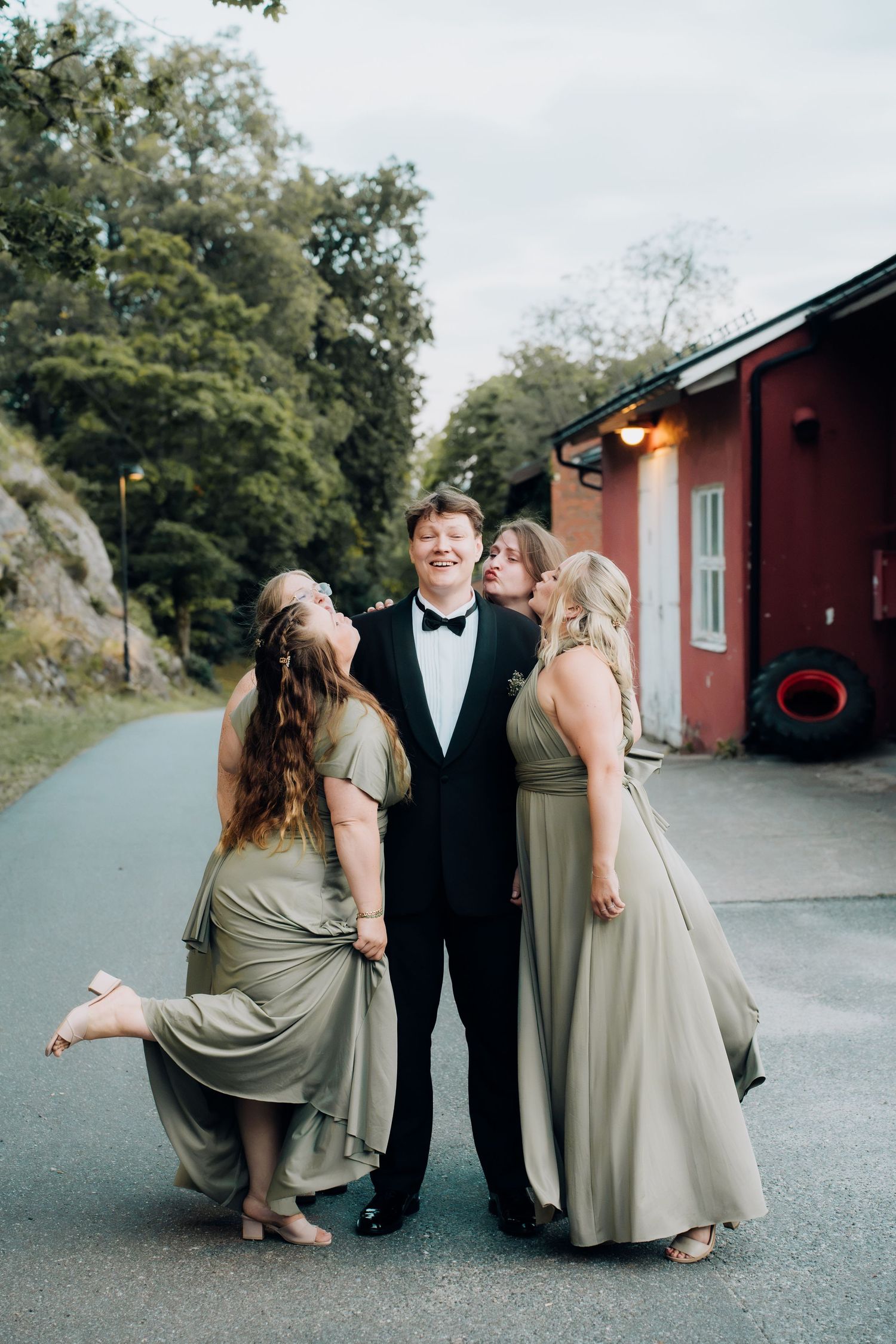 Groom in black tuxedo receives playful kisses from two bridesmaids in sage green dresses.