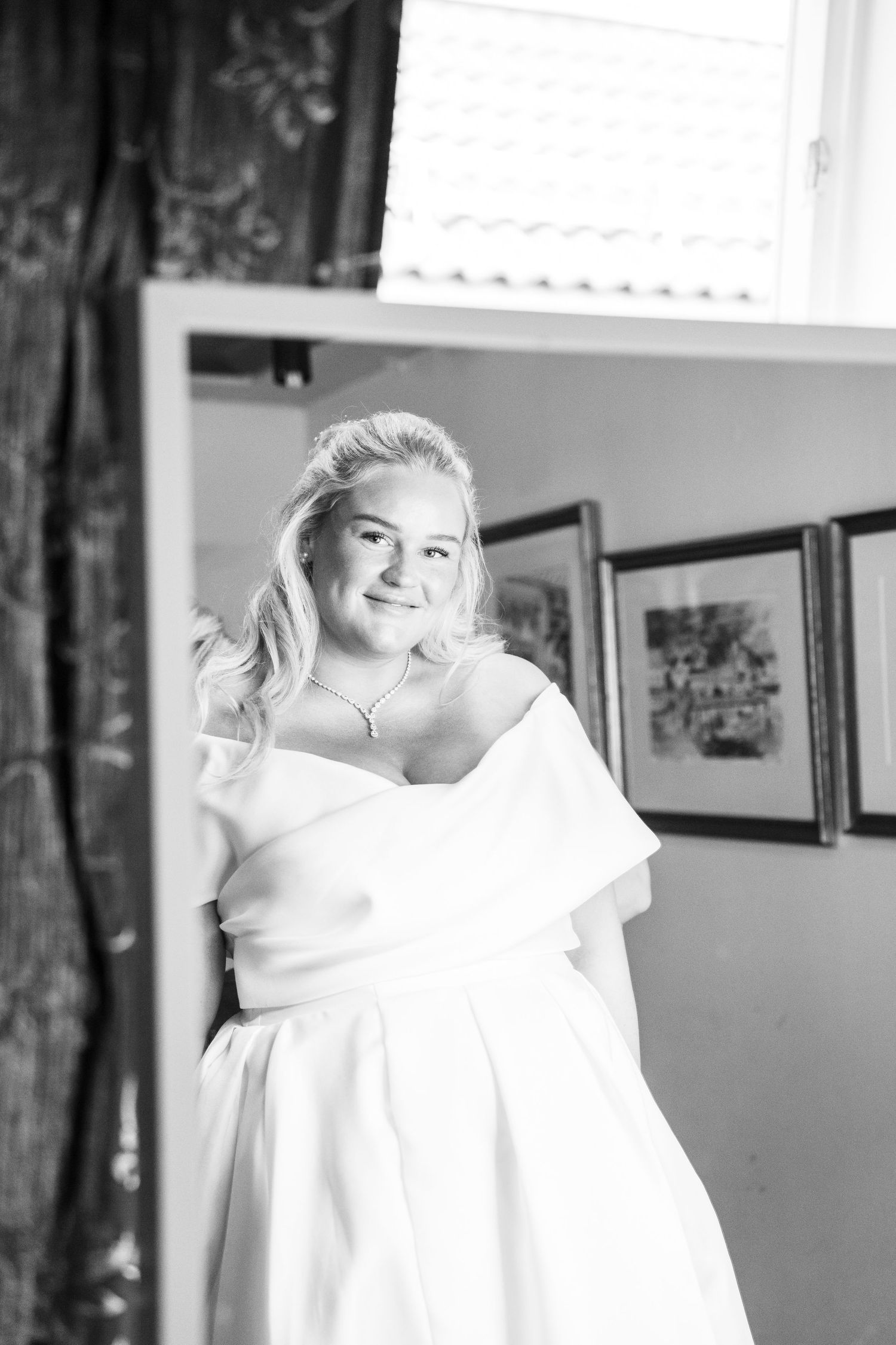 Black and white portrait of smiling bride in off-shoulder white gown reflected in mirror by window.