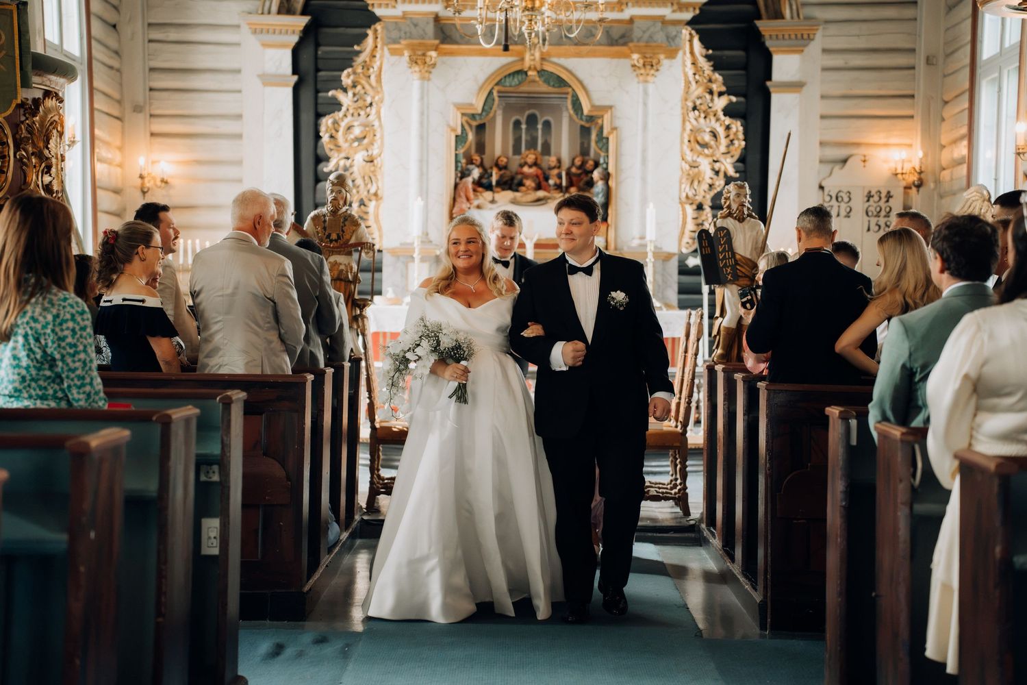 Bride and groom walk down church aisle after wedding ceremony, surrounded by seated guests in ornate historic church.
