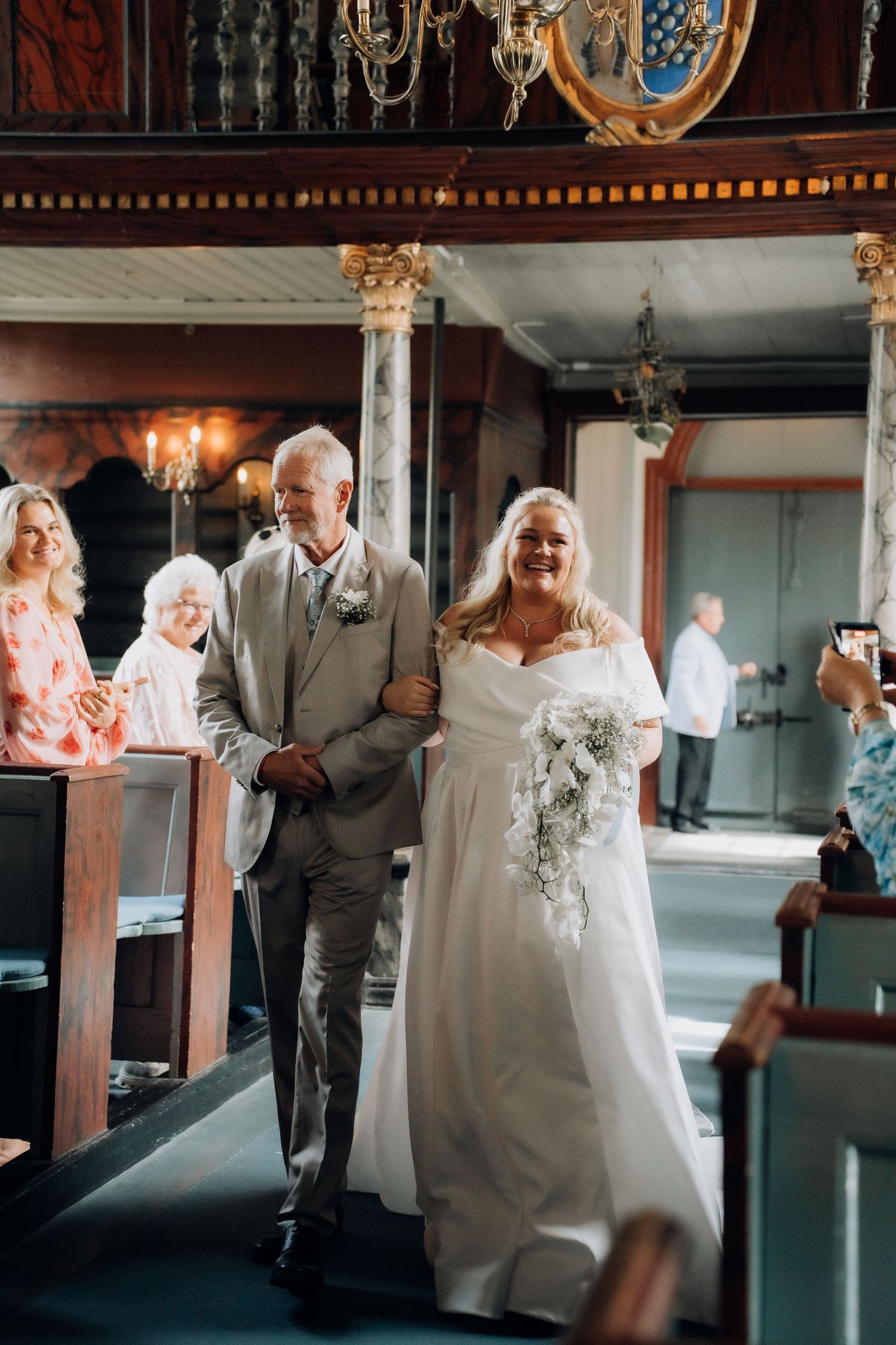 Bride in white off-shoulder gown with cascading bouquet walks down church aisle escorted by man in gray suit.