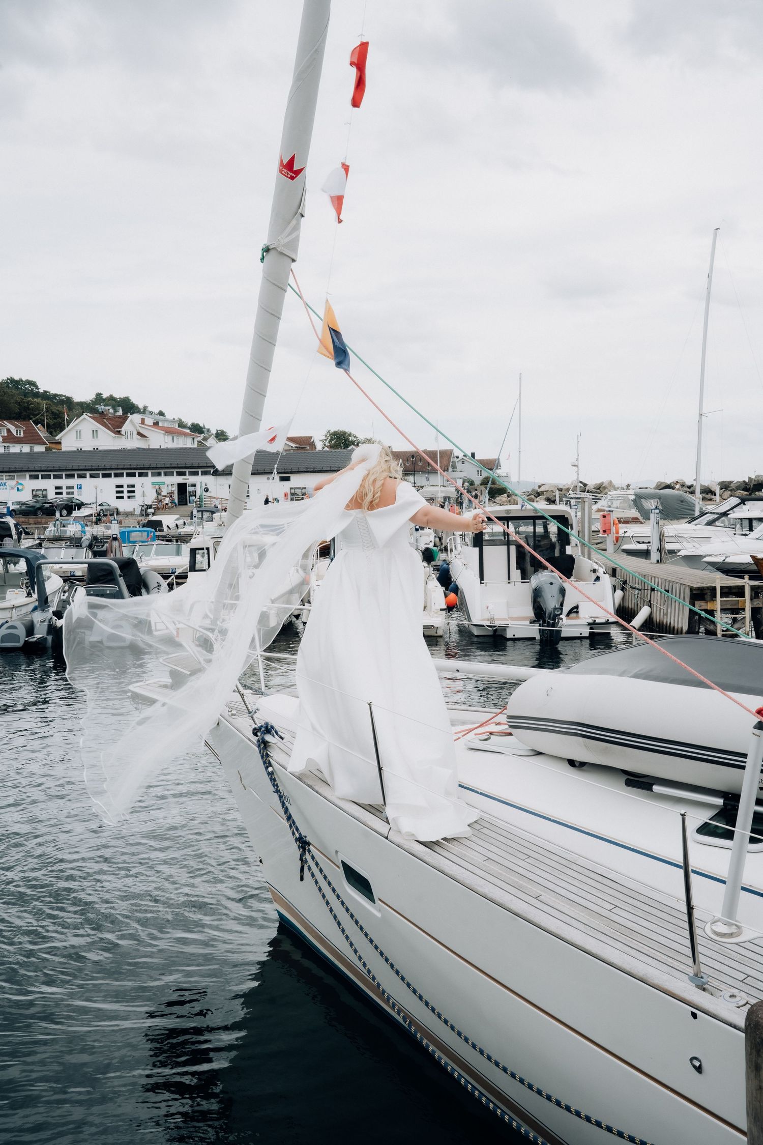 Bride in white gown standing on sailboat bow with veil flowing at a marina.