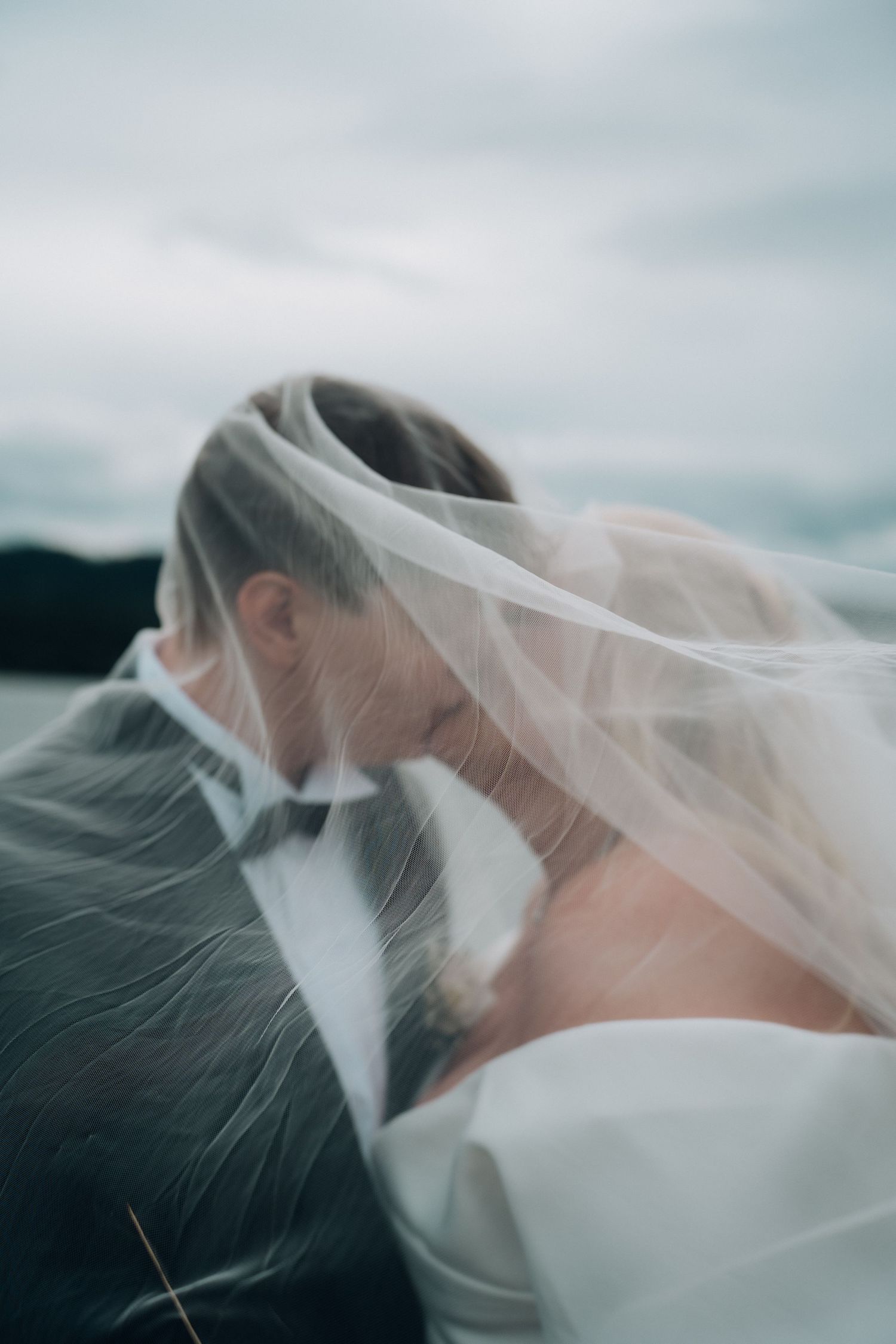 Bride and groom sharing a romantic kiss under a flowing veil against a cloudy sky backdrop.