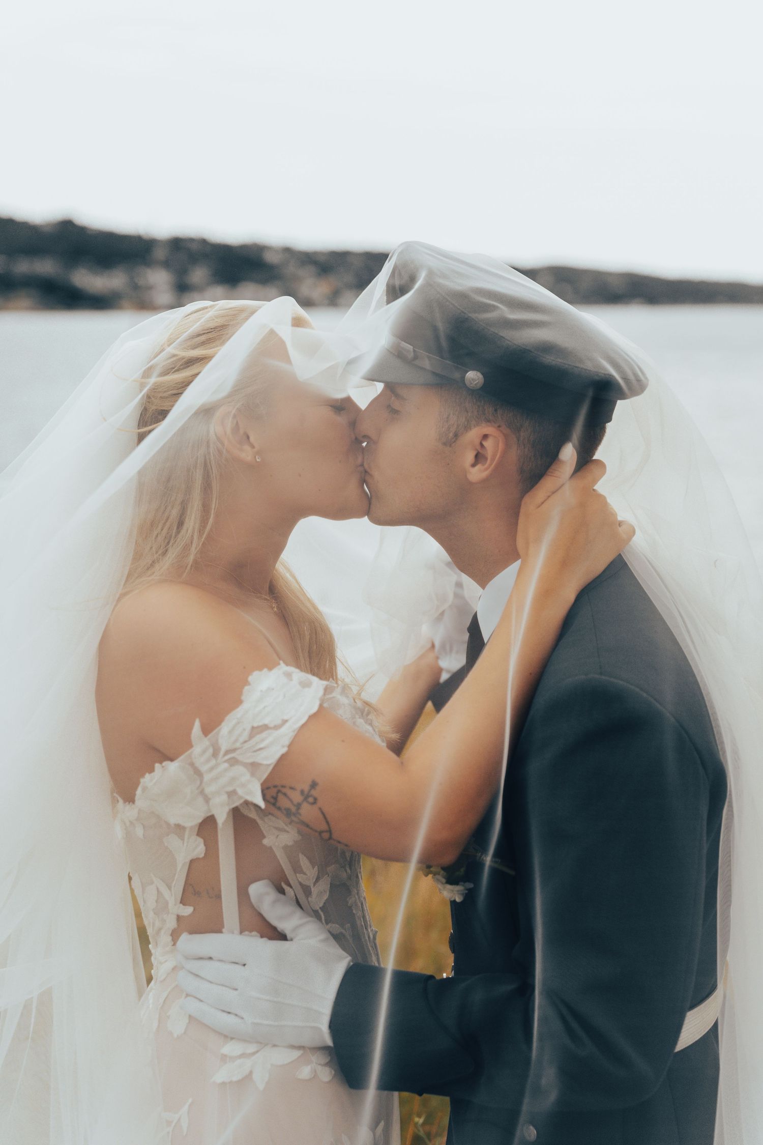 Bride and groom sharing romantic kiss under flowing veil by waterfront with groom in military uniform.