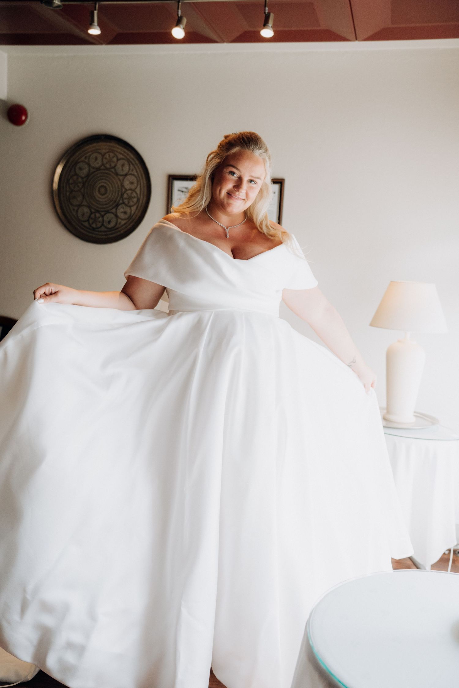 Smiling bride in off-shoulder white ball gown spreading her skirt wide in a bright hotel bridal suite.
