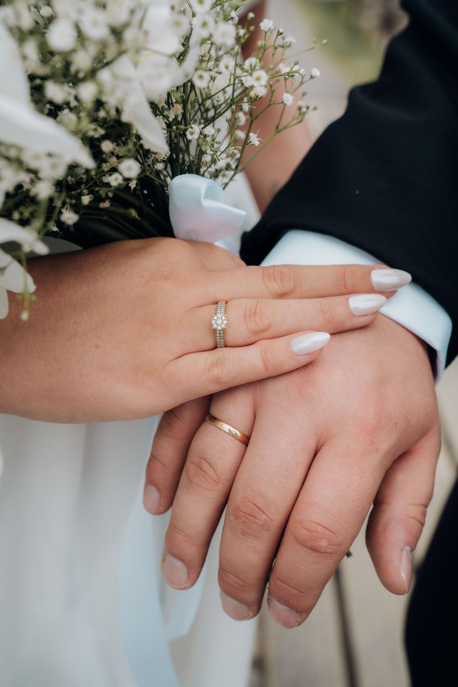 Close-up of newlyweds hands showing wedding rings and diamond engagement ring with white bouquet.
