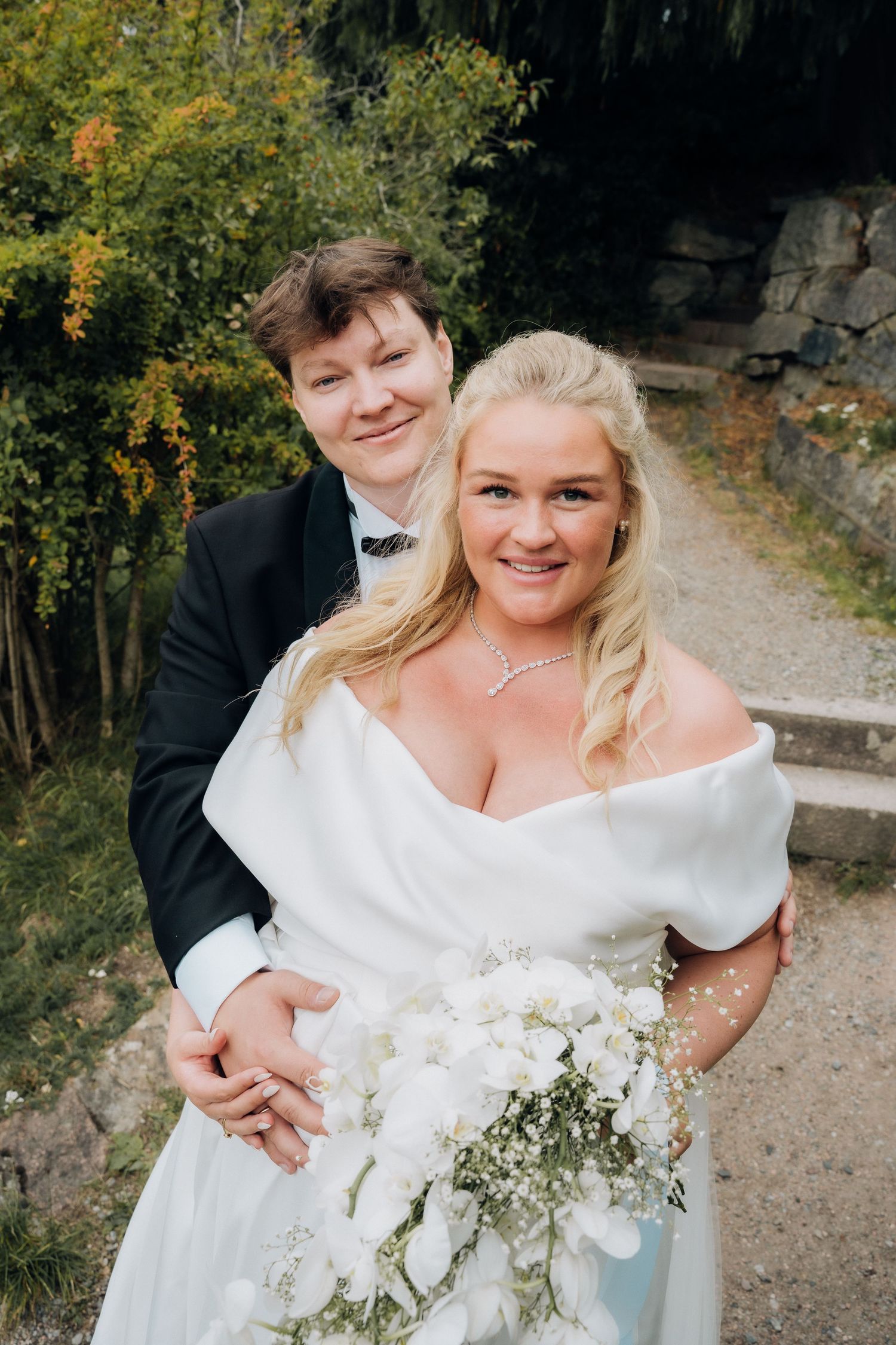 Smiling bride in white off-shoulder gown with white wrap and groom in black tuxedo posing outdoors.