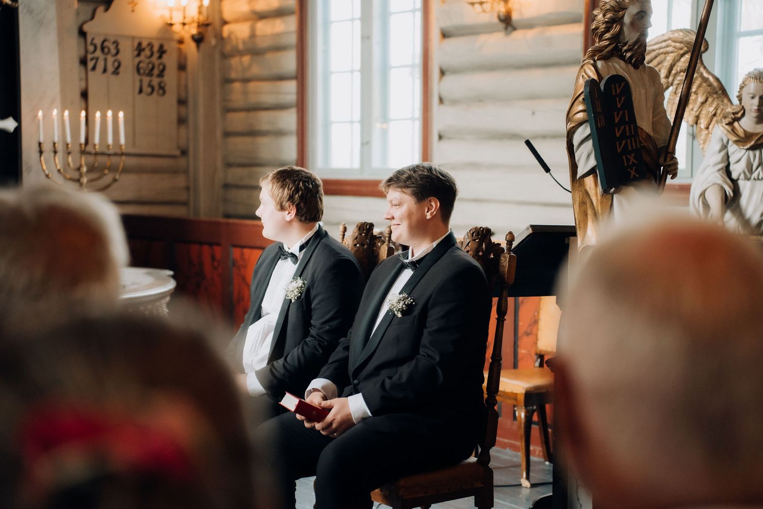 Two men in formal black tuxedos with bow ties seated during a wedding ceremony in a historic church.