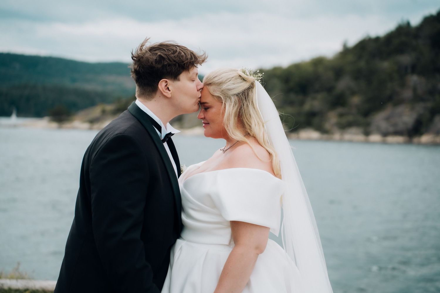Bride and groom sharing a tender kiss by a scenic waterfront on their wedding day.