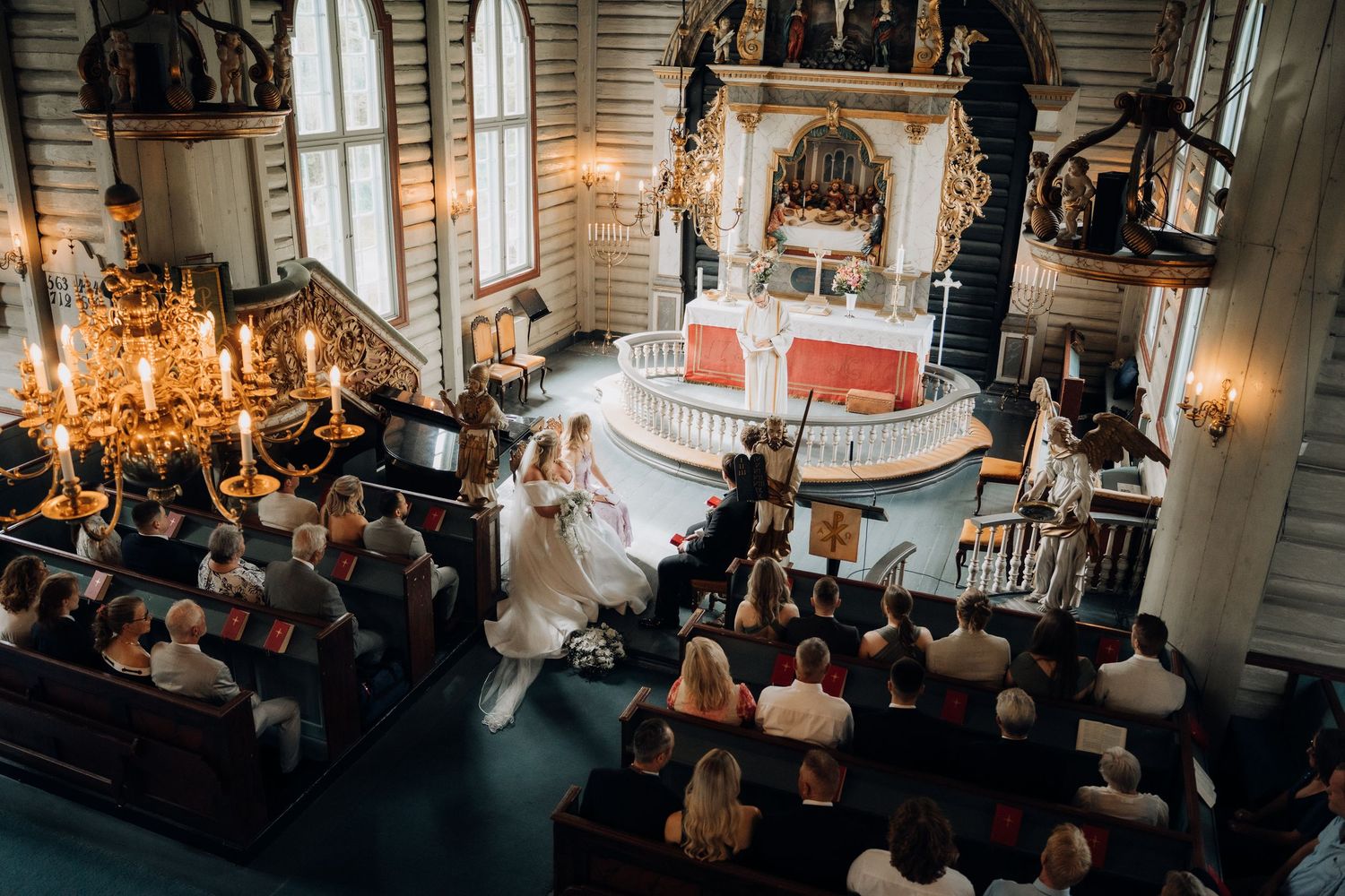 Aerial view of wedding ceremony in stunning historic church with chandeliers, wooden pews, and ornate baroque altar.