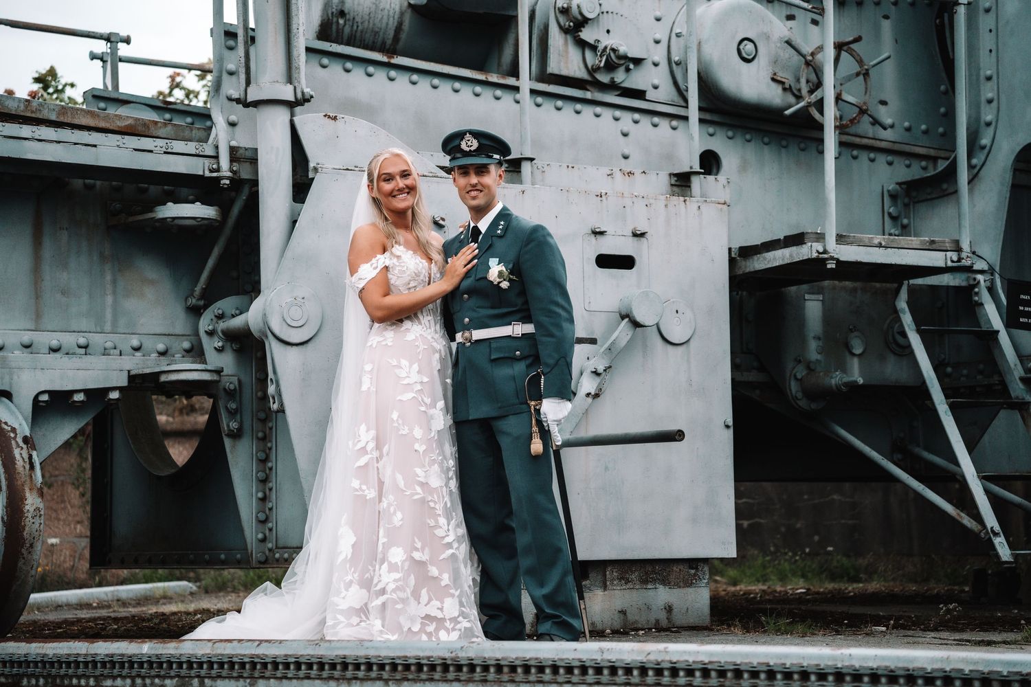 Blond brud i blondekjole med bare skuldre står sammen med brudgom i militæruniform ved siden av et veterantog.