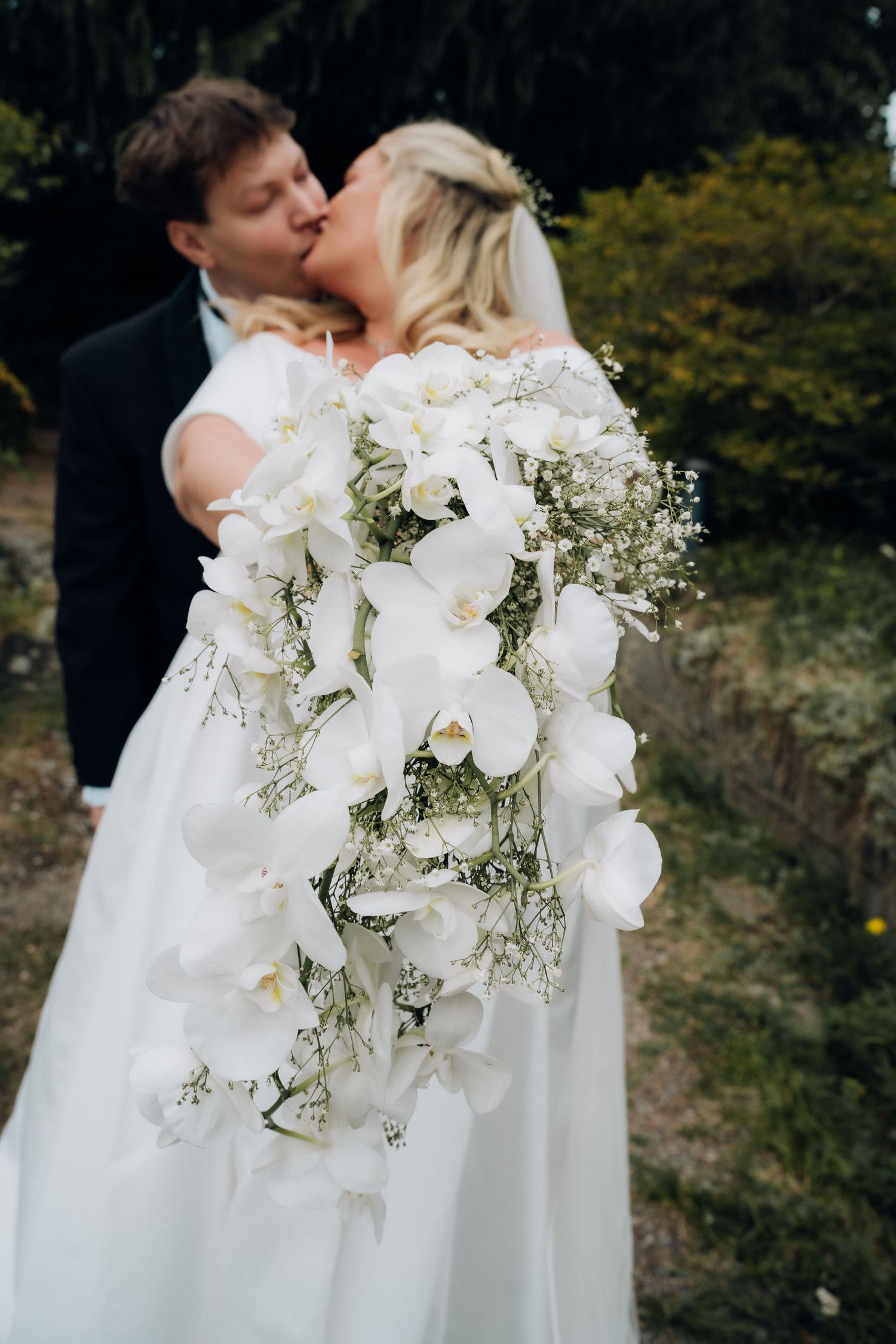 Bride and groom kissing behind a cascading white orchid and baby's breath wedding bouquet in a garden.