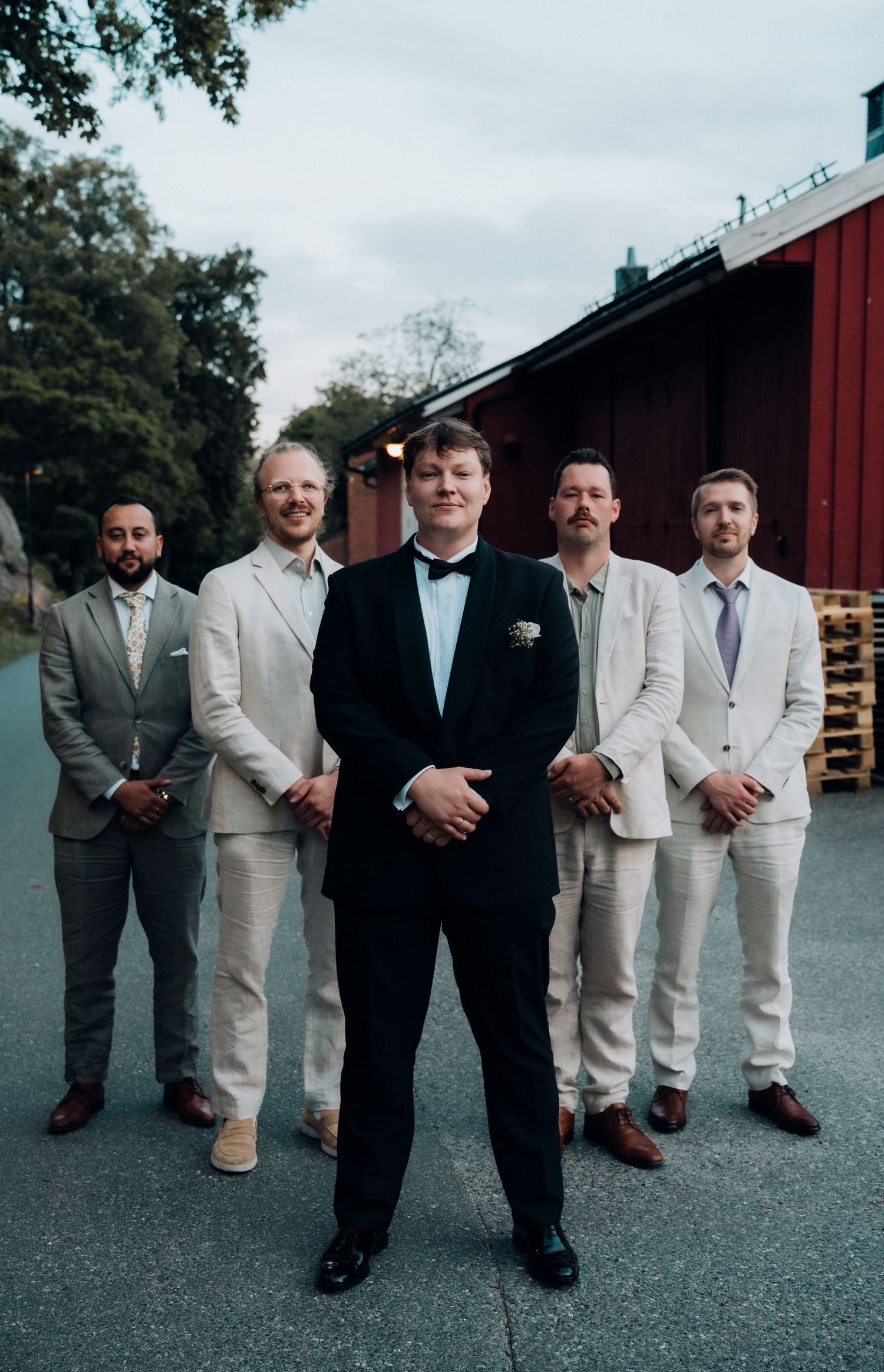 Groom in black tuxedo poses confidently with four groomsmen in linen suits at wedding.