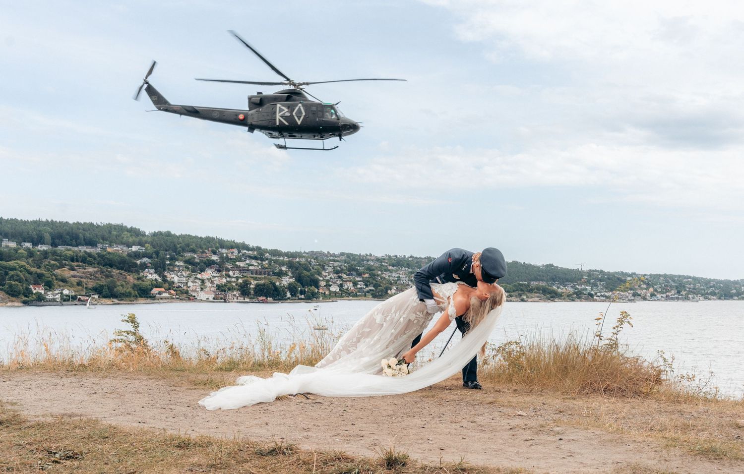 Brud og brudgom deler et romantisk kyss i en dukkende posisjon på stranden mens et militærhelikopter flyr over dem.