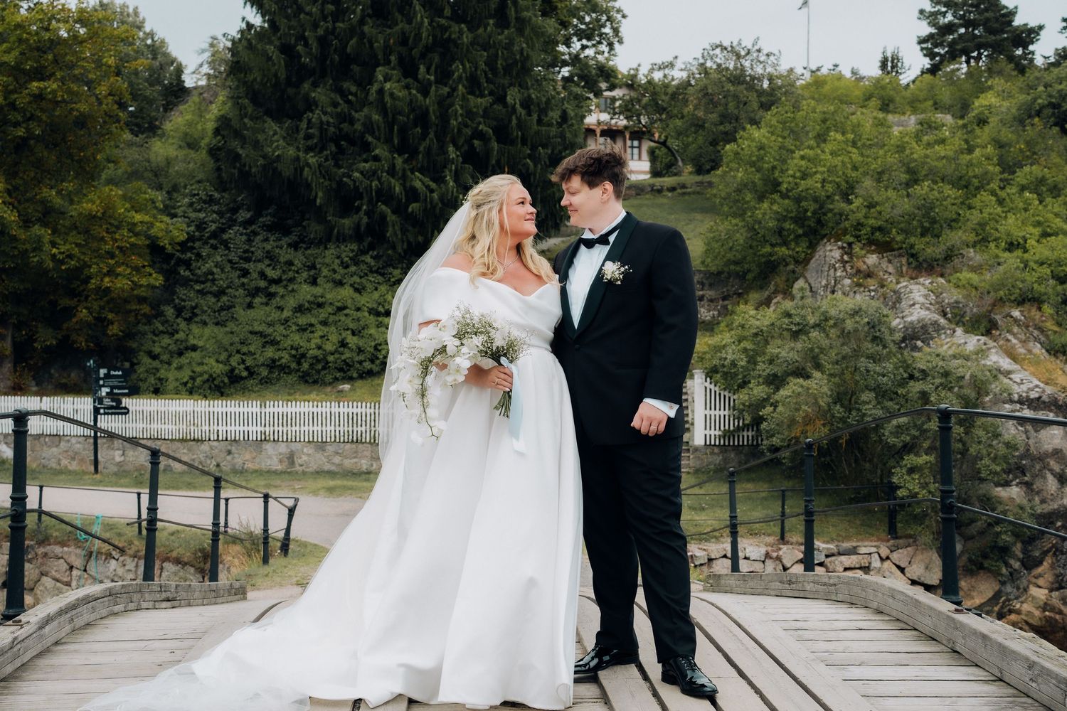 Bride and groom smiling at each other on a wooden bridge surrounded by lush green scenery.