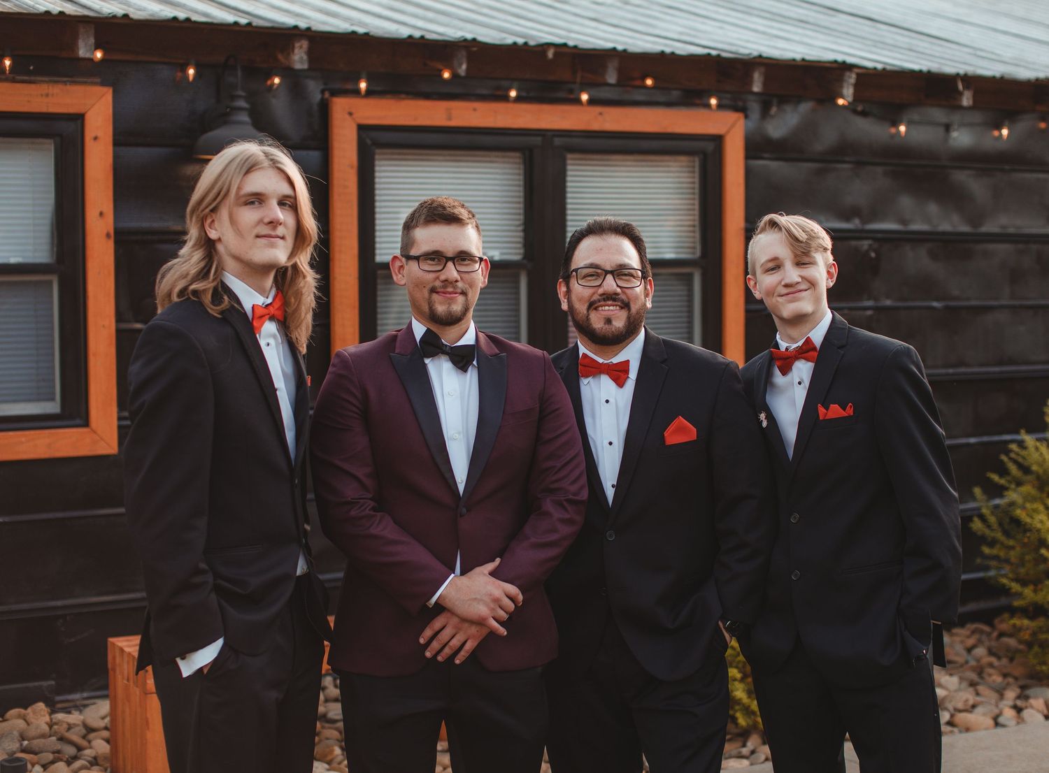 Groom in a burgundy tuxedo standing with his groomsmen outside a dark wood barn