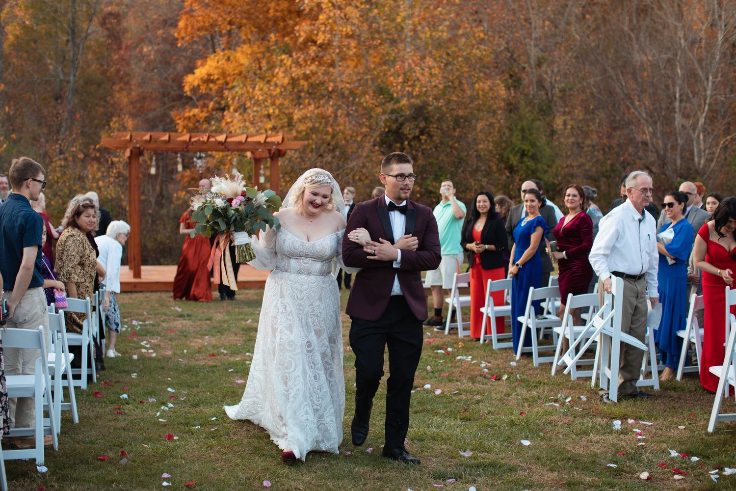 Bride escorted down the aisle toward a wooden pergola with fall foliage behind