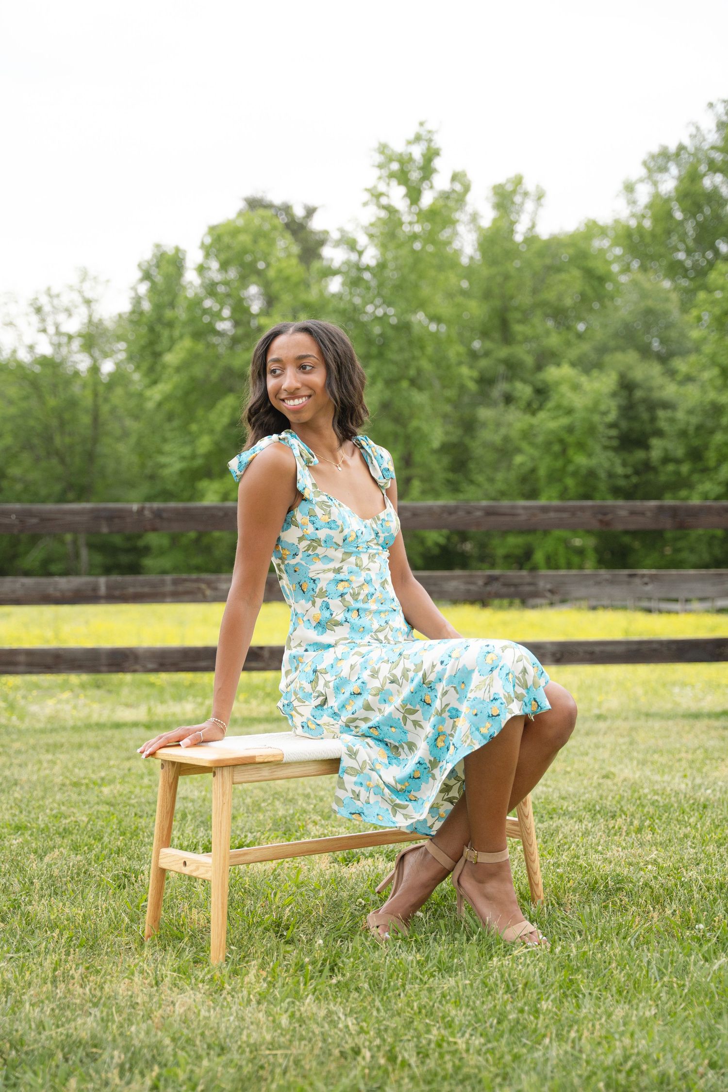 Graduate in a floral dress seated on a stool in a pasture with trees behind her