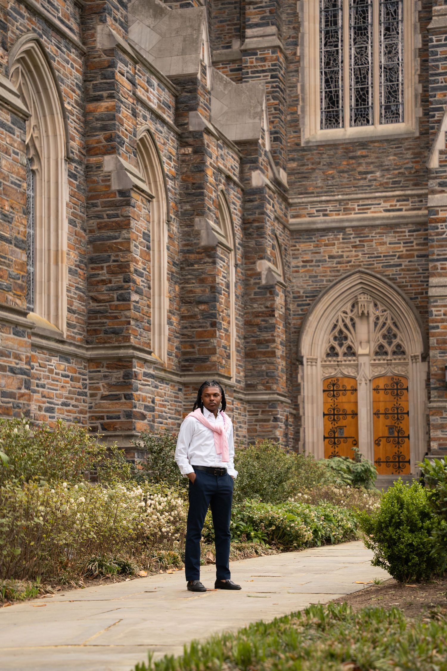Graduate posing on a garden path beside the stone walls of a Gothic chapel