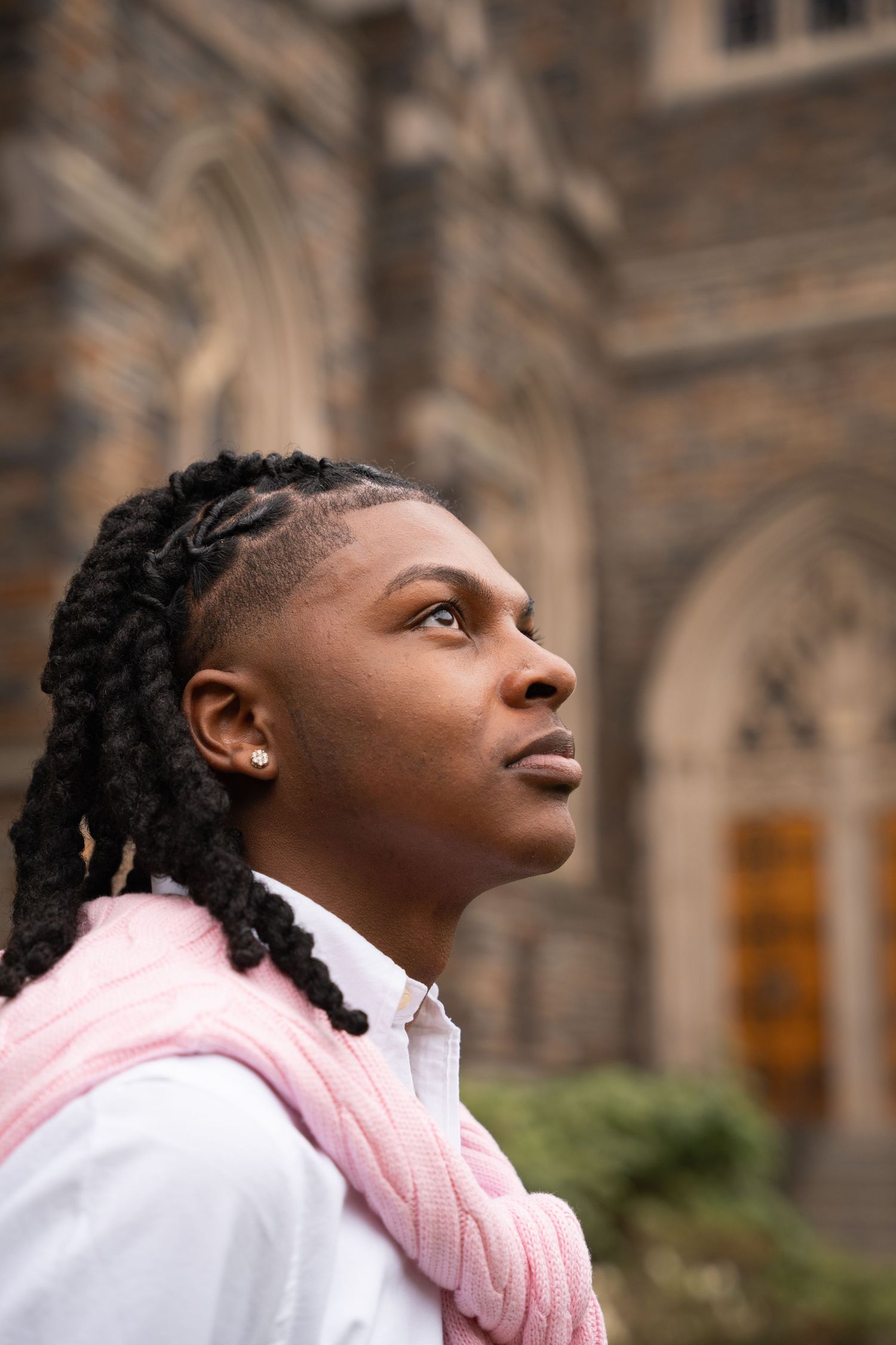 Graduate looking upward with a pink sweater draped over his shoulders by a stone chapel
