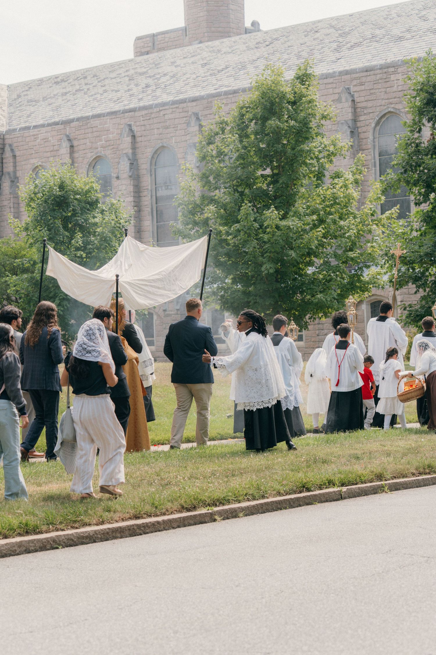Corpus Christi procession with altar servers and a white canopy outside a brick Catholic church
