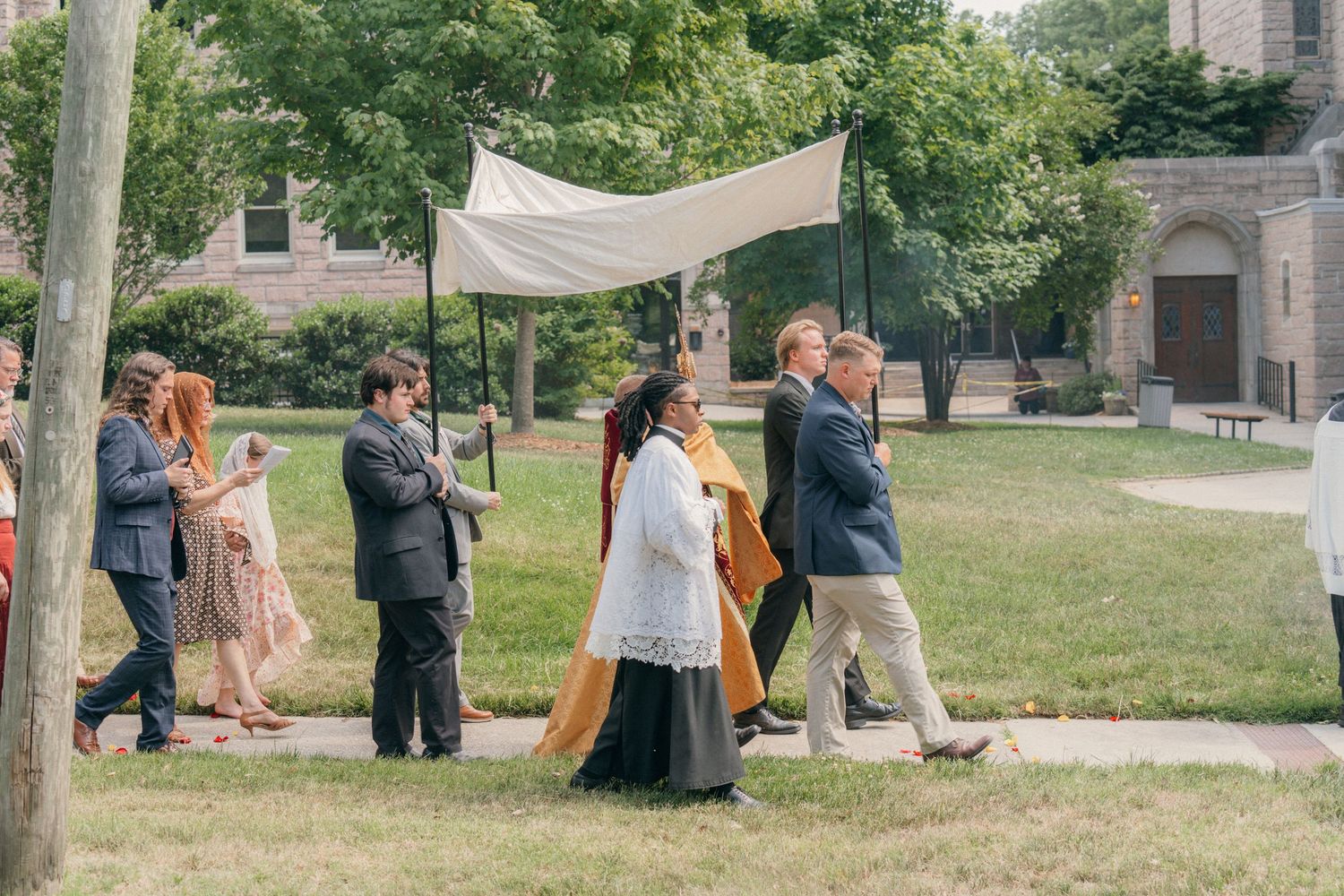 Corpus Christi Eucharistic procession walking under a white canopy across a church lawn
