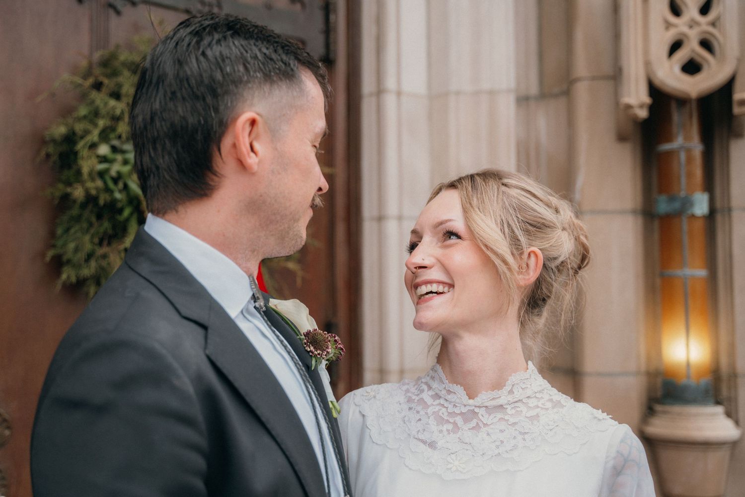Bride and groom smiling at each other outside a stone church doorway on their wedding day
