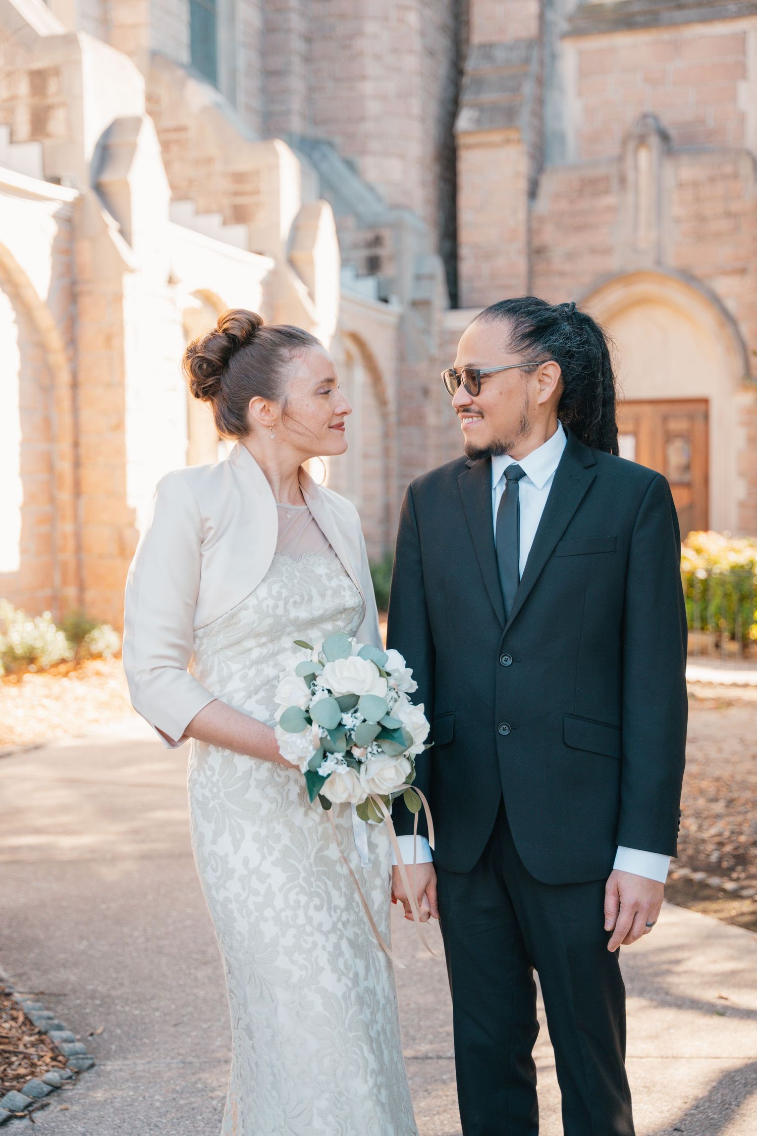 Bride with a white bouquet and groom in a black suit outside a brick church