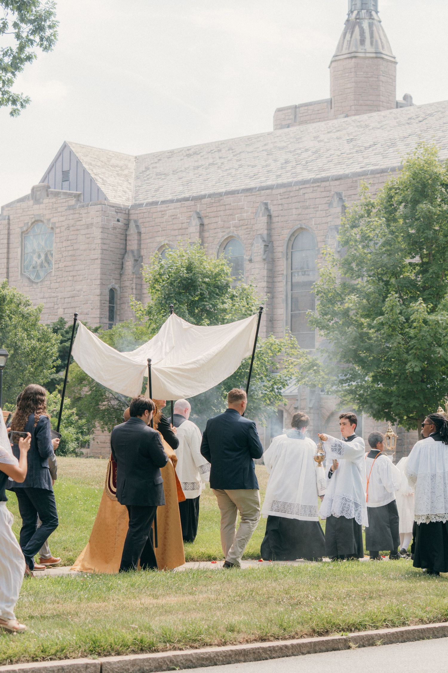 Parishioners and altar servers gather around the canopy in a Corpus Christi procession outside a brick church