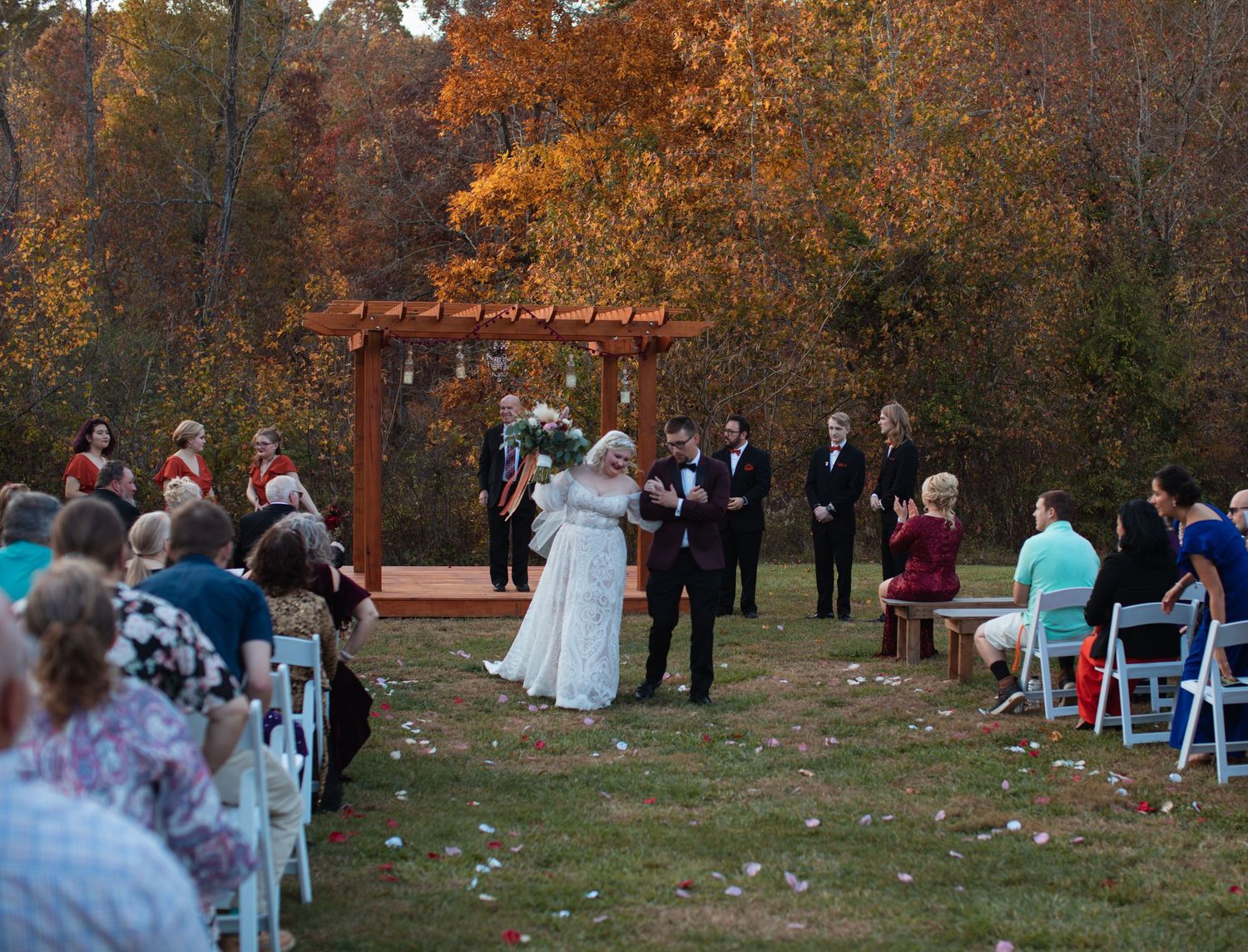 Wide view of an outdoor wedding ceremony under a pergola surrounded by autumn trees