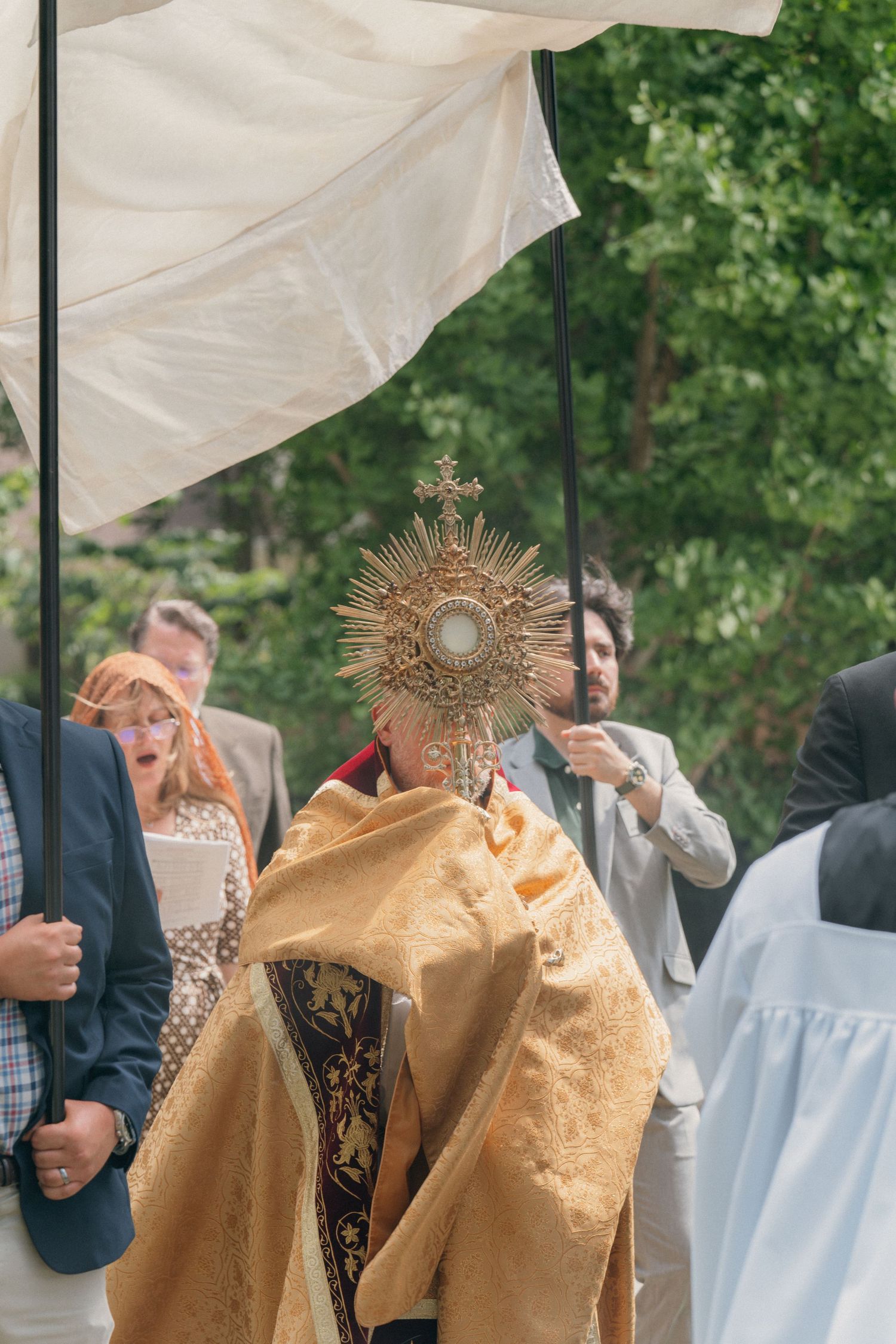 Priest in a gold humeral veil carrying the Blessed Sacrament under a canopy, parishioners behind