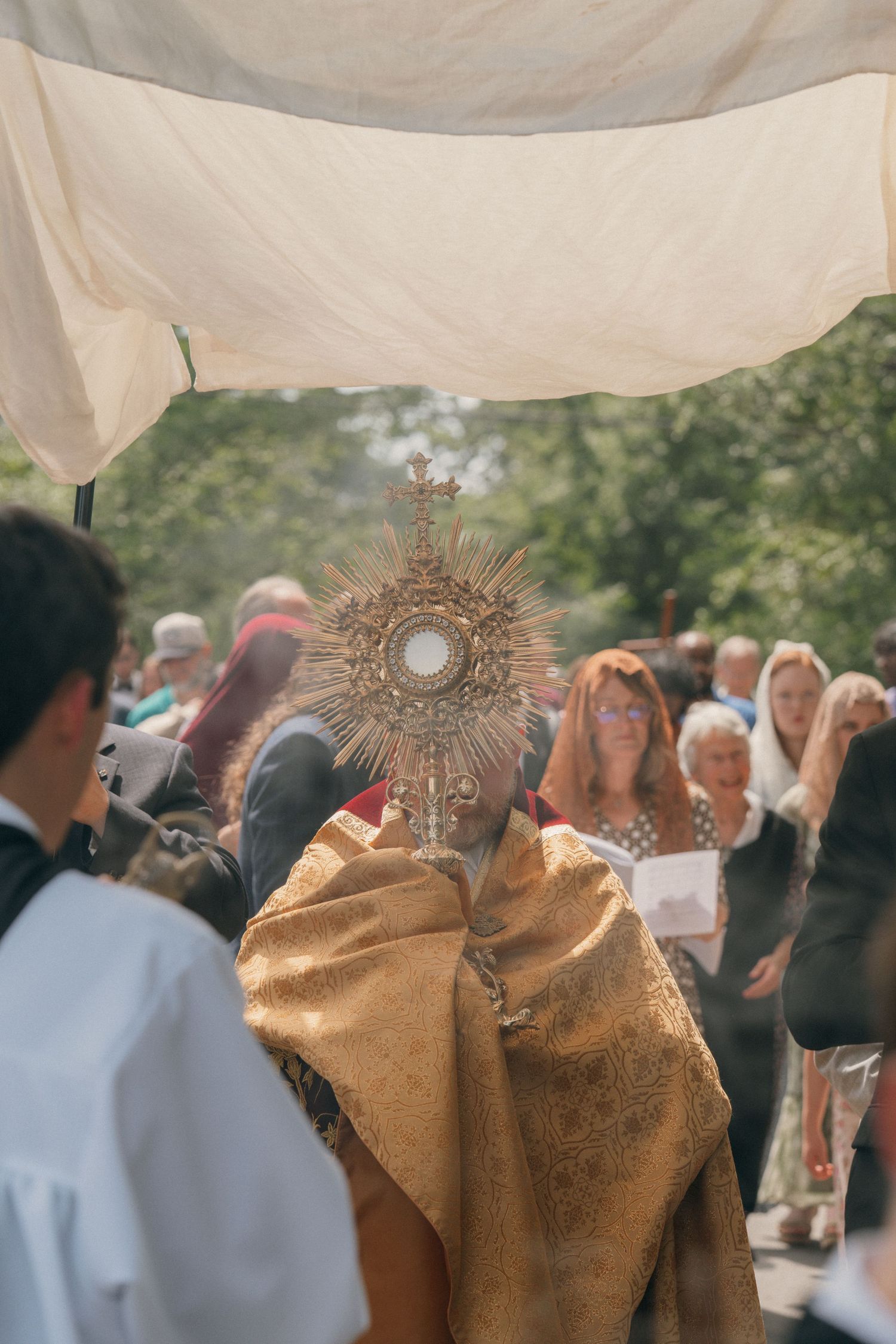 Priest holding the monstrance under a canopy as a crowd of parishioners looks on during Corpus Christi