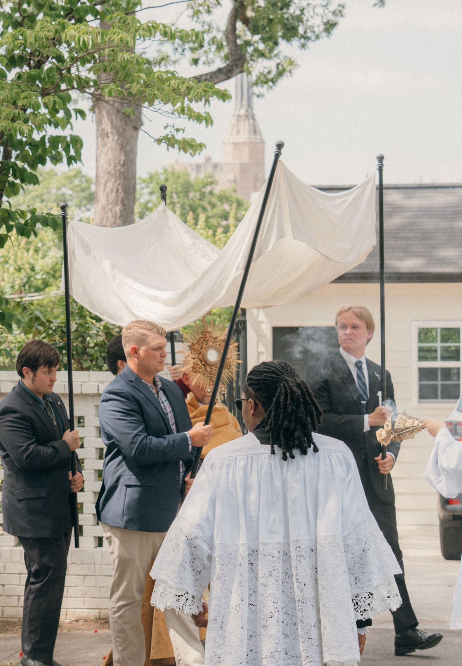 Canopy bearers and a priest walking past a white house in a Corpus Christi procession