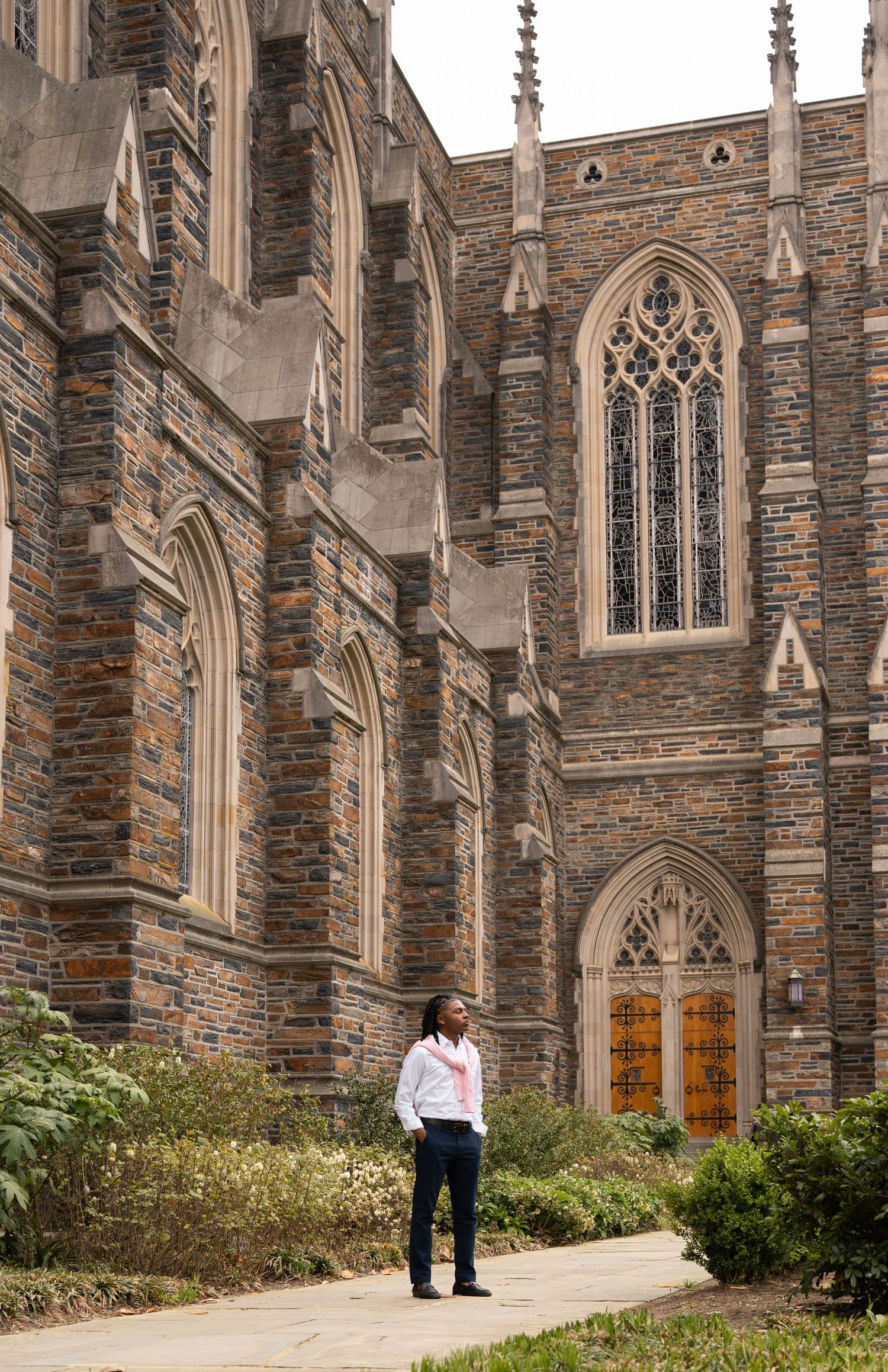 Wide shot of a graduate standing on a garden path below a towering Gothic chapel