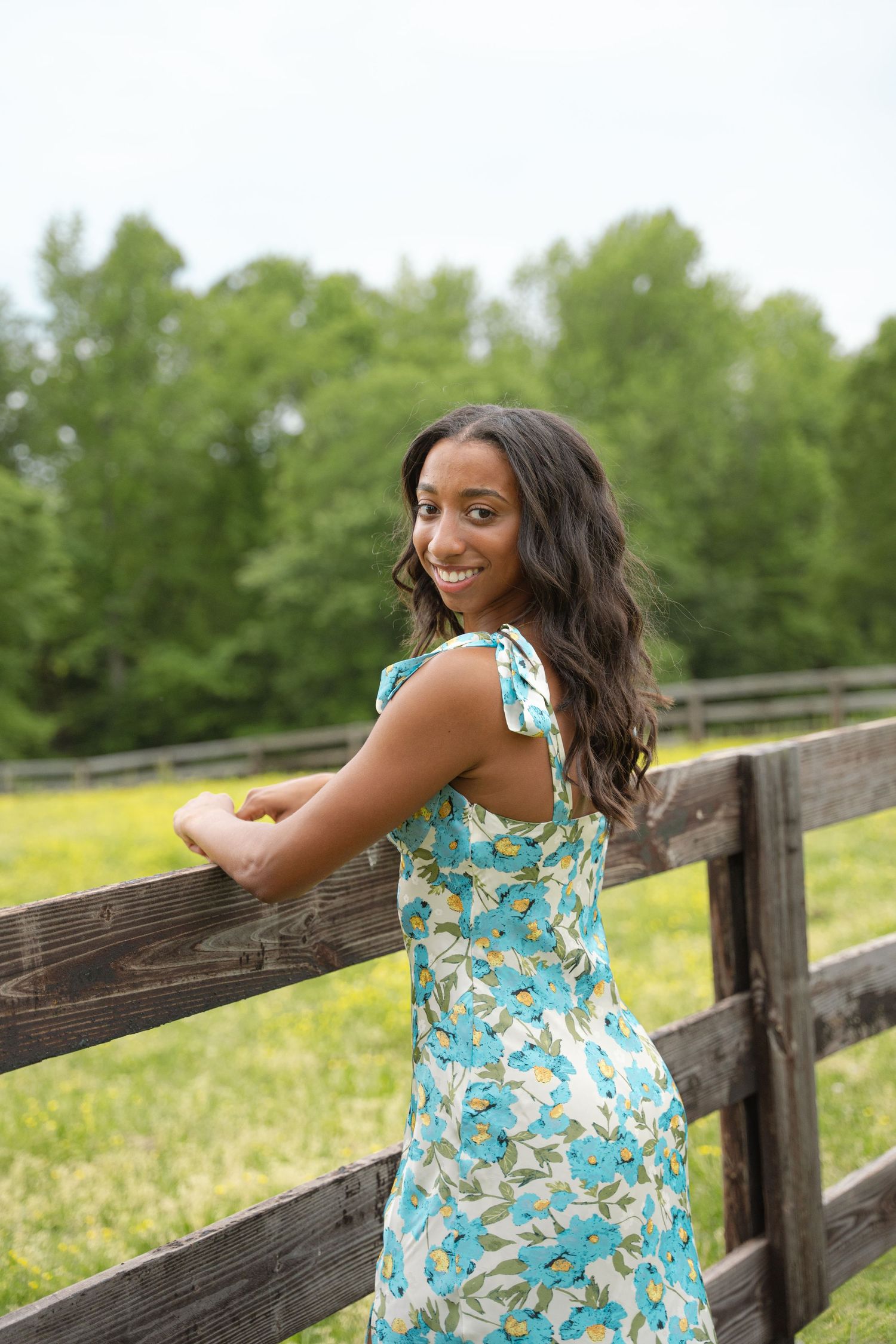 Graduate in a blue floral sundress leaning on a split-rail fence in a green pasture