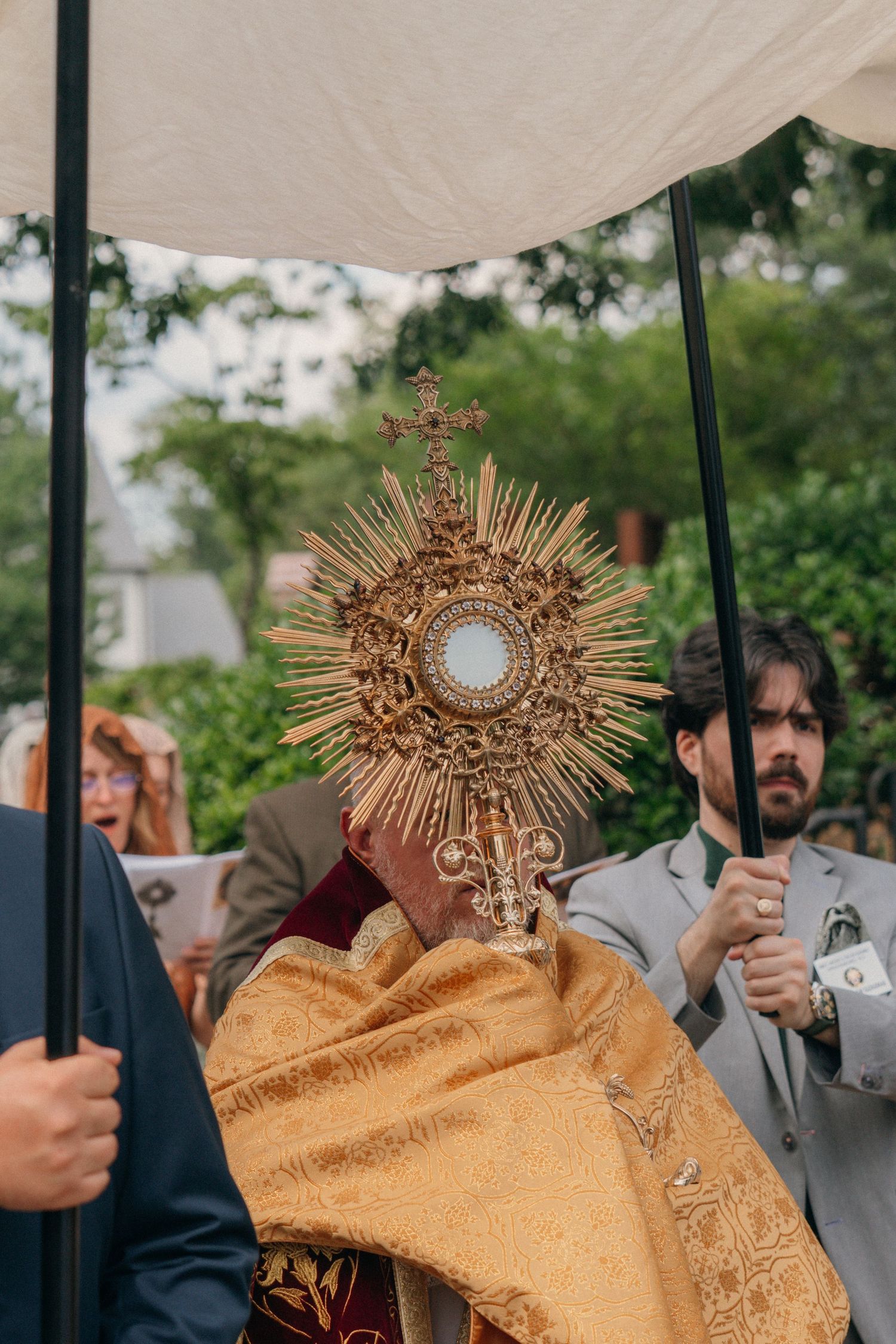 Men holding canopy poles beside the priest carrying the monstrance in a Corpus Christi procession